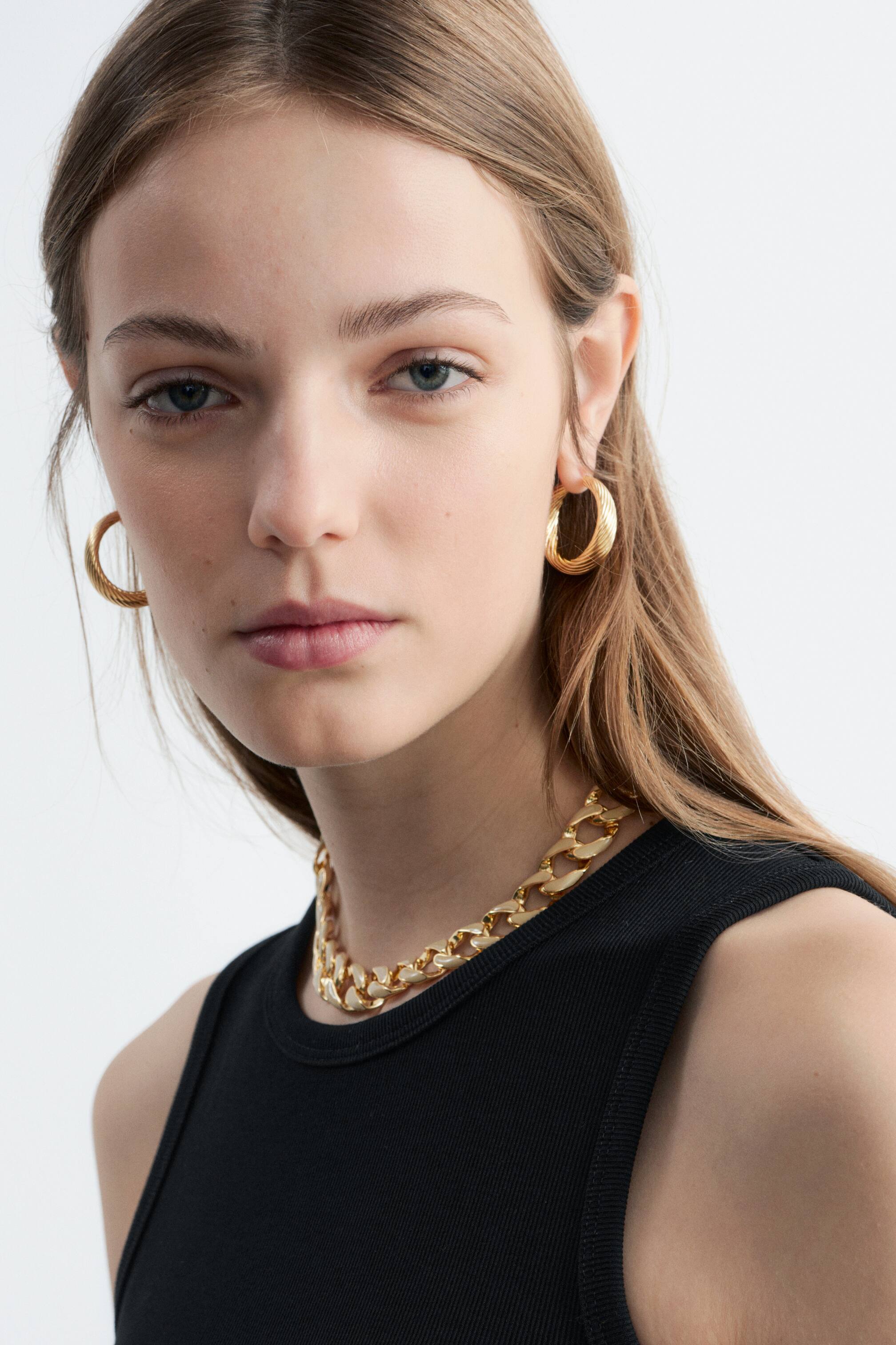 Visualizza immagine più grande: Orecchini a cerchio con texture in tonalità dorata sono indossati su entrambe le orecchie. Una collana a catena con maglie spesse in tonalità dorata è adagiata sopra un top nero a coste.