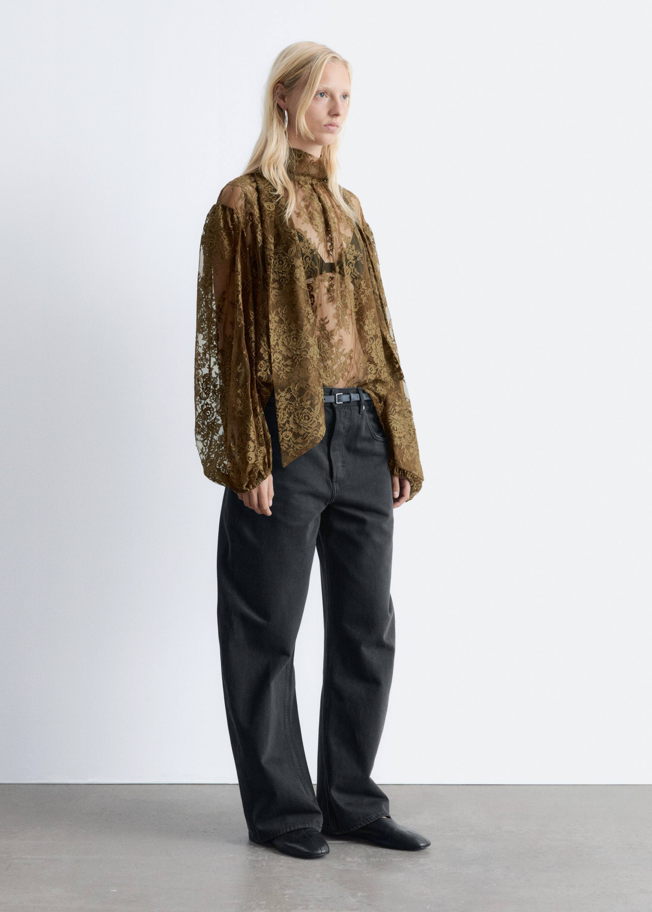 Ingrandisci l'immagine: Una blusa di pizzo trasparente verde oliva, con collo alto e maniche lunghe e voluminose con polsini arricciati, viene presentata da un'angolazione laterale, rivolta a destra. La blusa è stratificata su un reggiseno scuro e infilata in pantaloni ampi grigio scuro, fissati con una sottile cintura nera.