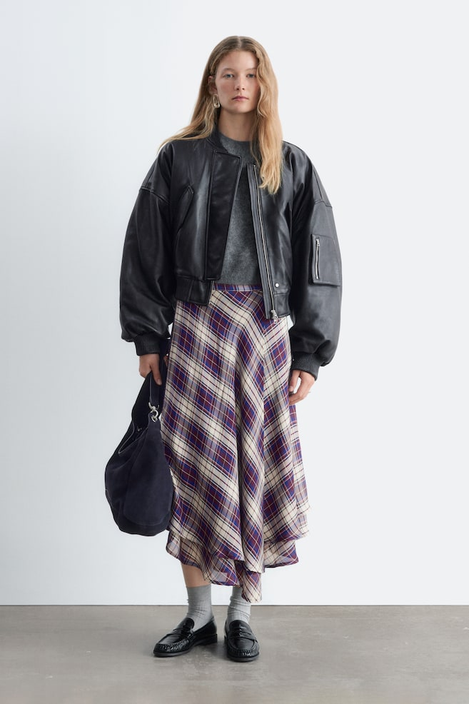 A black cropped leather bomber jacket is worn in a front-facing view, showcasing an oversized silhouette with dropped shoulders and ribbed-knit trim at the collar, cuffs, and hem. It is layered over a grey top to highlight its cropped waist length and features a silver-tone front zip alongside a zipped utility pocket on the sleeve. - 1