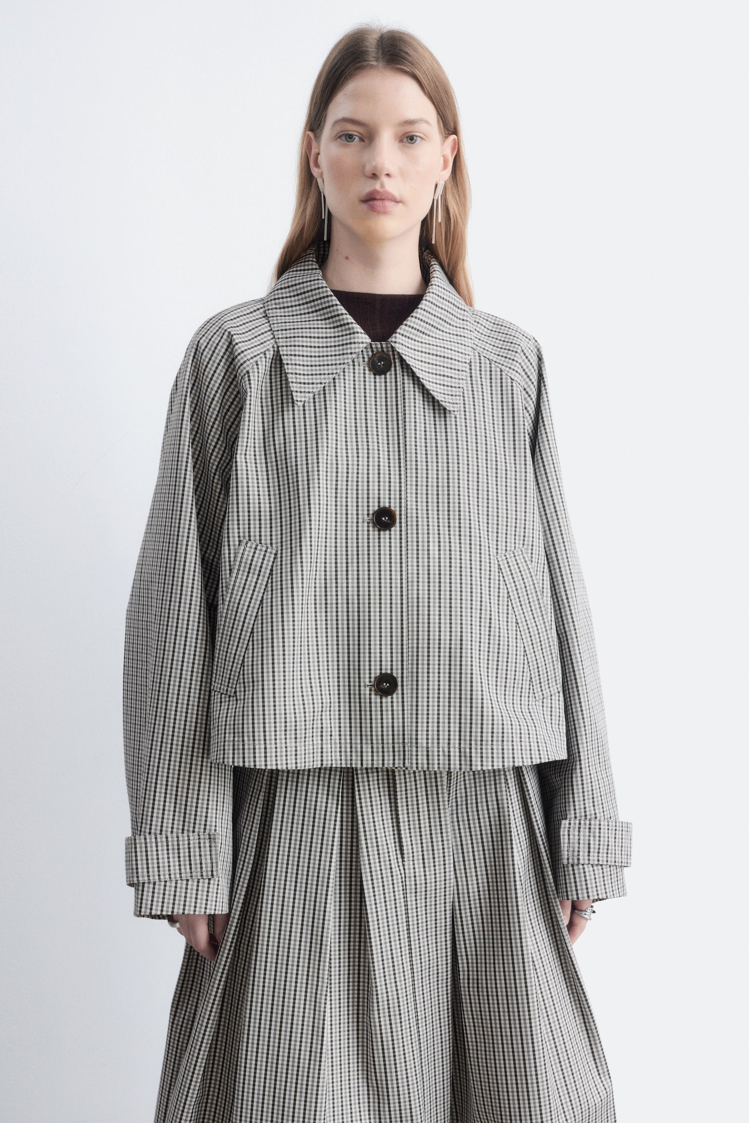 Une veste courte prÃ©sentant un petit motif Ã  carreaux noir et blanc, un col pointu et de grands boutons foncÃ©s sur le devant est portÃ©e par le modÃ¨le. La veste est associÃ©e Ã  une jupe plissÃ©e assortie et superposÃ©e Ã  un haut foncÃ© Ã  col montant. - 1