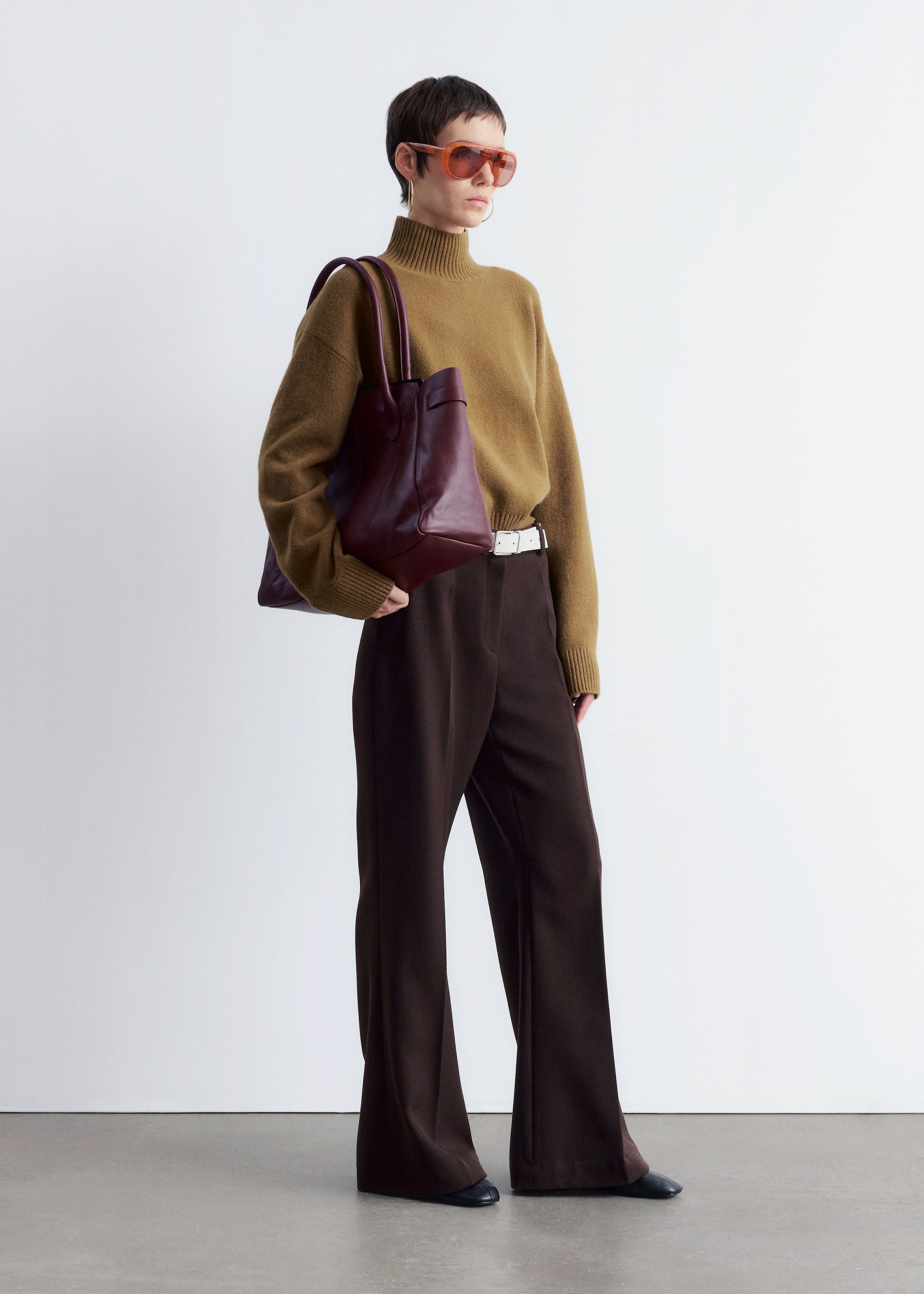 Agrandir l'image: Une tenue complète est présentée, mettant en avant un pull à col roulé olive-brun ample avec des poignets côtelés et un pantalon large marron foncé, resserré à la taille par une fine ceinture blanche. Un grand sac fourre-tout en cuir bordeaux profond est porté, et le look est accessoirisé avec des lunettes de soleil teintées orange, des boucles d'oreilles en or et des chaussures plates foncées.