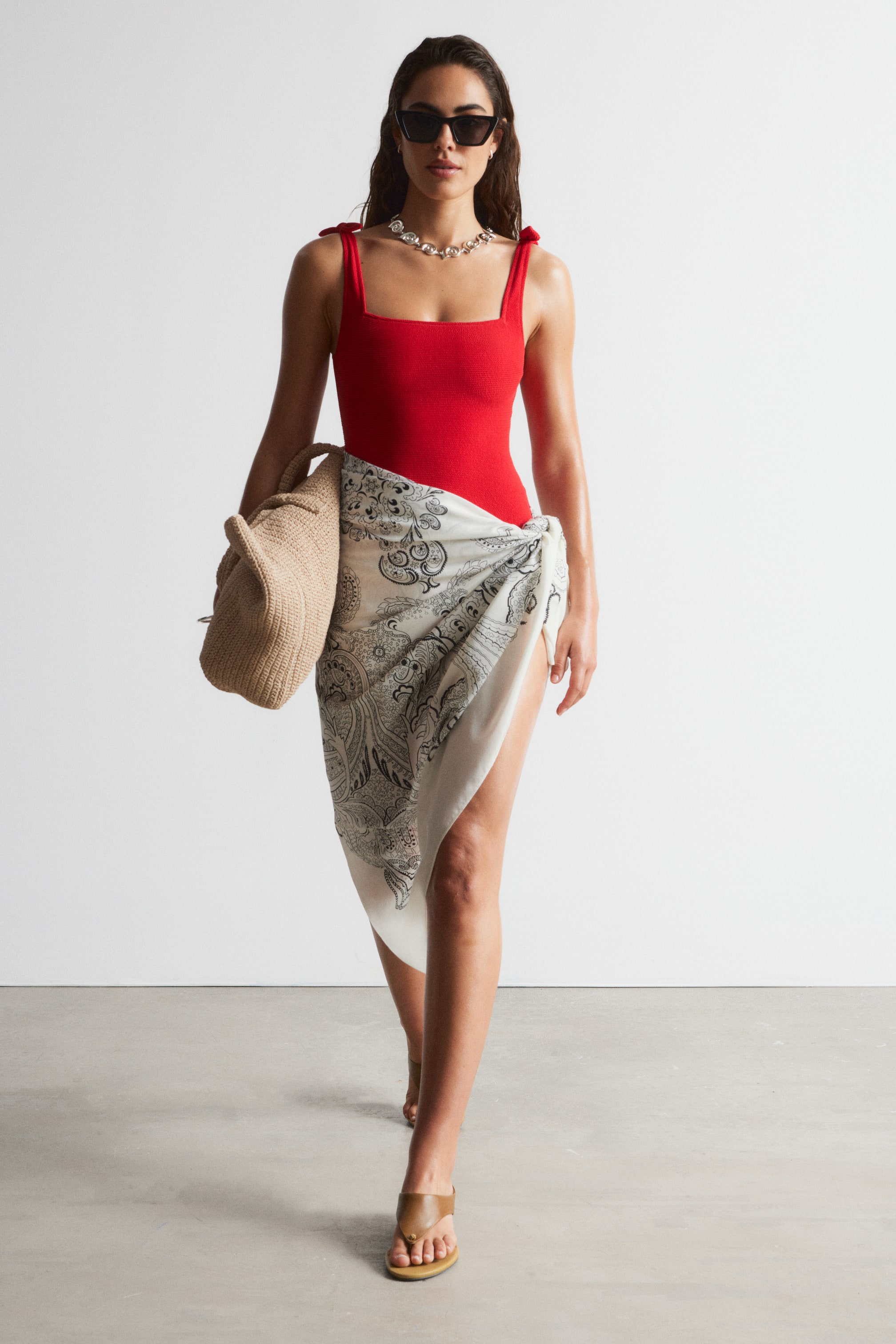 View larger image: The model is wearing a red swimming costume with a square neckline and tied shoulder straps. She has paired it with a white paisley sarong, tied elegantly over the costume. Completing the look, she carries a woven straw bag and is accessorised with sunglasses, a necklace, and sandals.