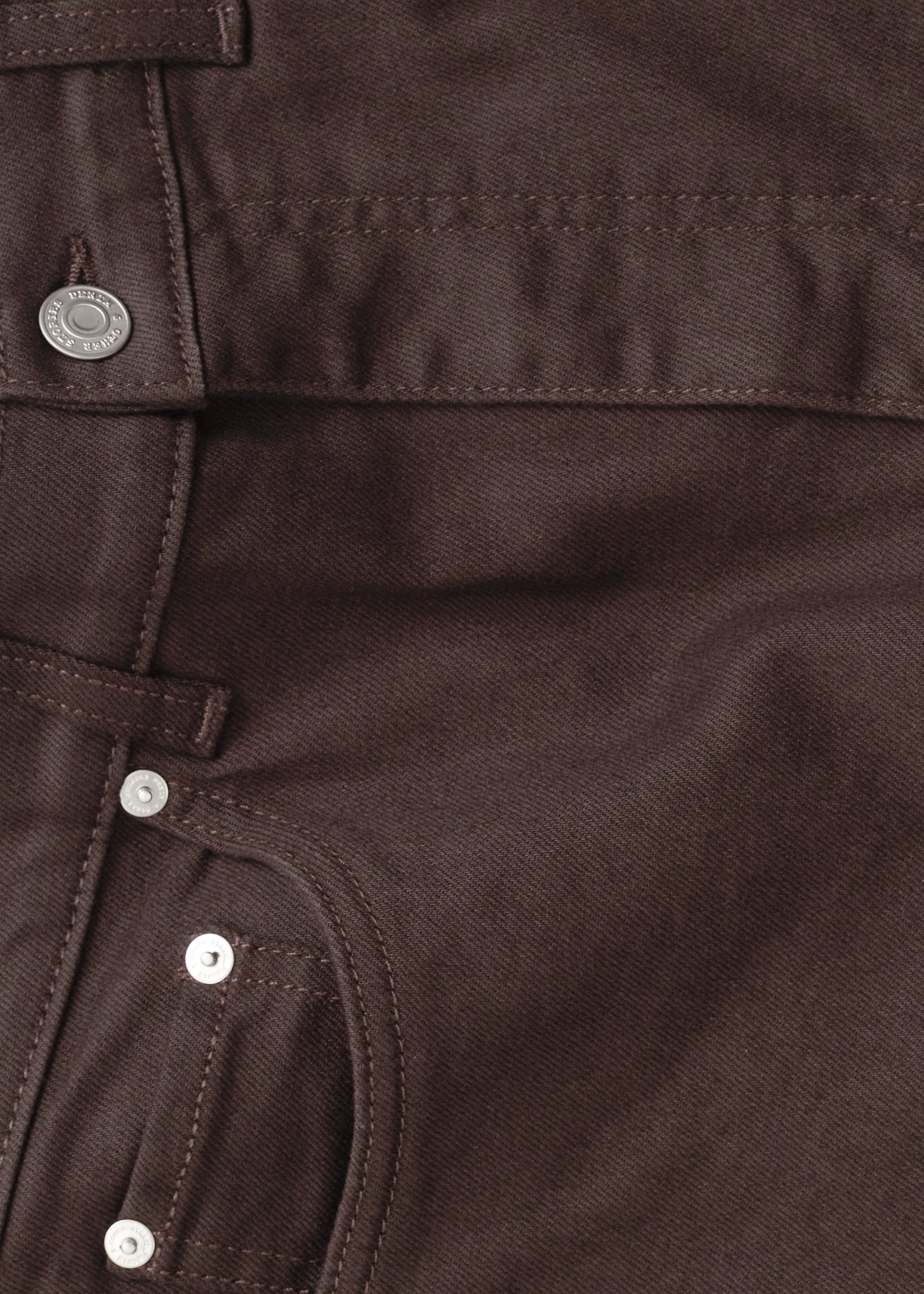 Se større billede: Et par mørkebrune højtaljede jeans ses fra en top-down vinkel, der viser deres øverste front. Jeansene har en sølvfarvet hovedknap, en lynlås med synlige syninger og en forlomme med små sølvnitter, karakteristisk for et 5-lomme denimdesign, med bæltestropper synlige langs toppen.