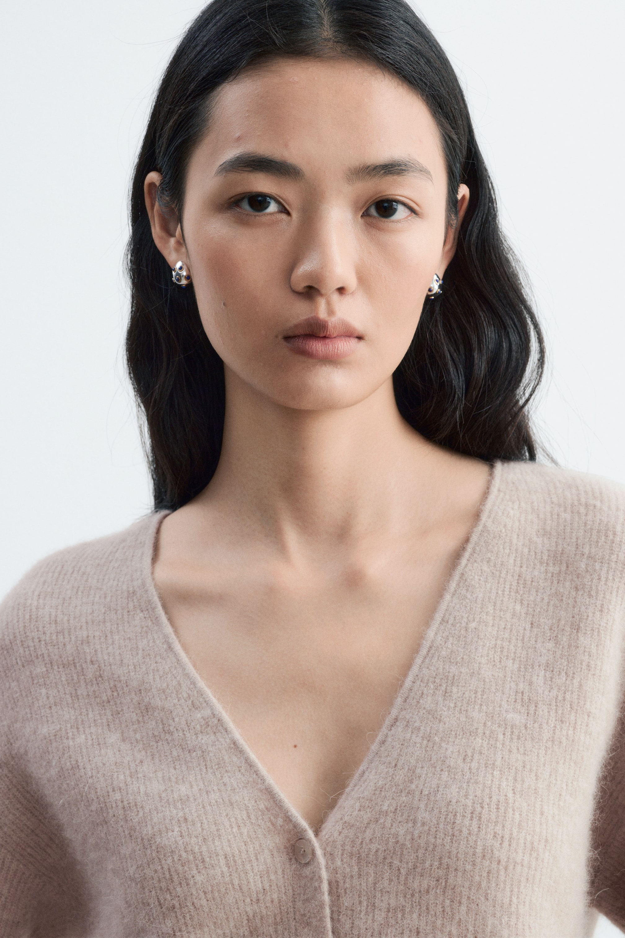 Vezi imaginea mai mare: Un cardigan bej deschis, din tricot cu model striat, cu decolteu în V și închidere cu nasturi, este purtat de model. Sunt purtați și cercei metalici, lucioși, rotunzi, cu pietre închise la culoare, care completează decolteul curat al cardiganului.