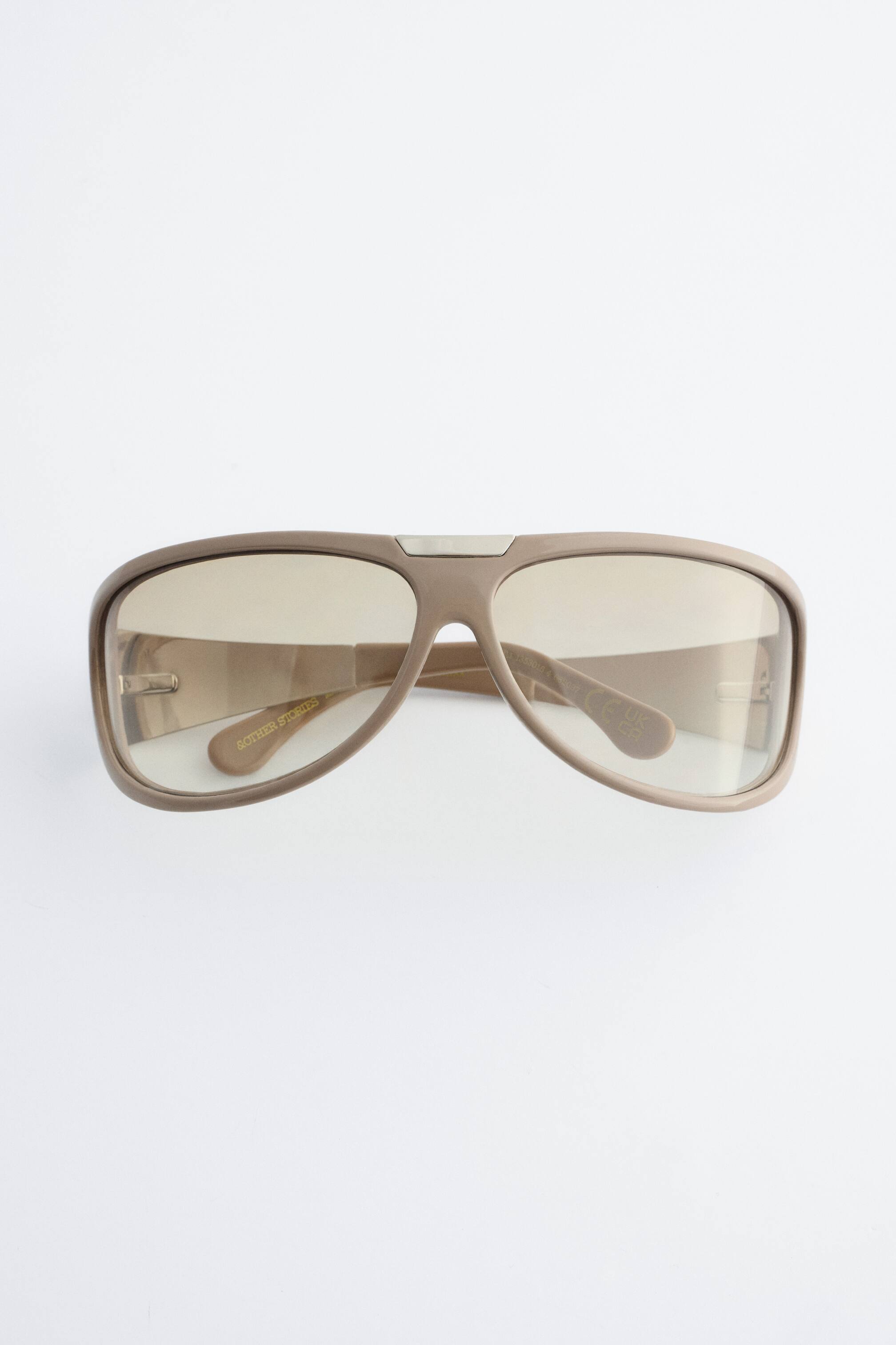 Agrandir l'image: Des lunettes de soleil ovales sont présentées de face, avec une monture épaisse couleur taupe et de grands verres teintés brun clair, arrondis. Le design met en avant un petit détail métallique plat argenté sur le pont supérieur, tandis que les branches repliées sont visibles à travers les verres.