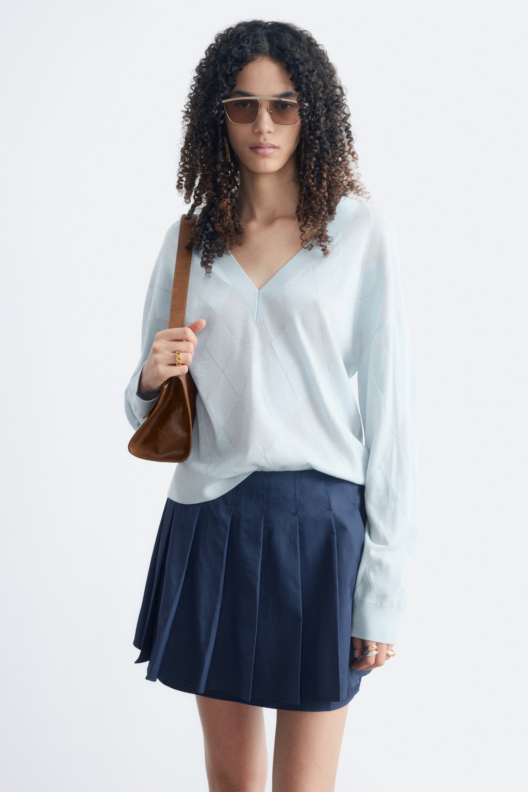 View larger image: A light blue V-neck jumper is on display, featuring a subtle diamond pattern across the front and long sleeves. It is paired with a navy pleated mini skirt, shown from the front.