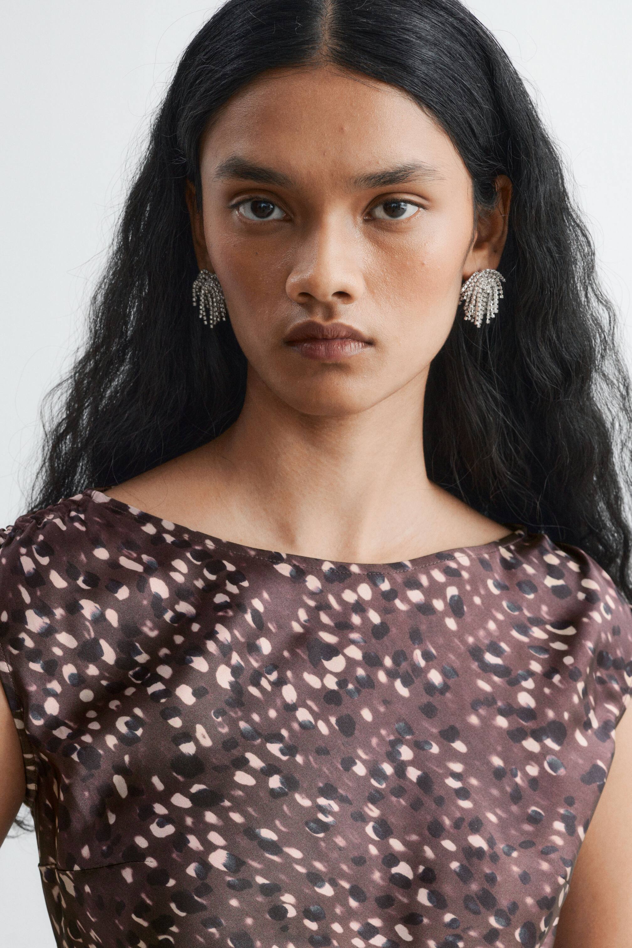 View larger image: Silver-toned fringe earrings embellished with rhinestones are being worn by the model, paired with a brown and beige patterned satin dress. The dress features a high round neckline and subtle gathers at the shoulders.
