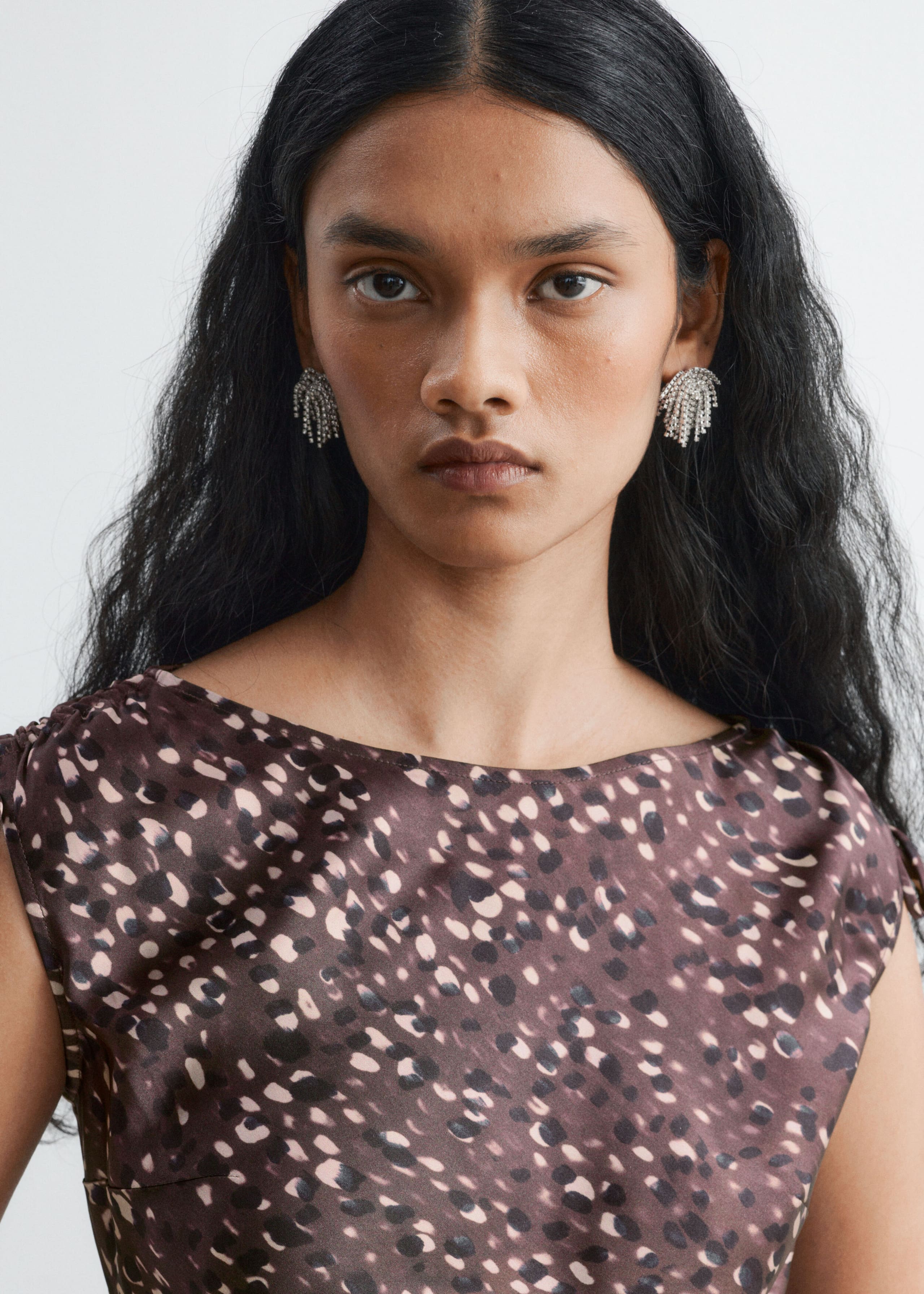 Silver-tone fringe earrings embellished with rhinestones are worn by the model and paired with a brown and beige patterned satin dress. The dress features a high crew neckline and subtle gathers at the shoulders.
