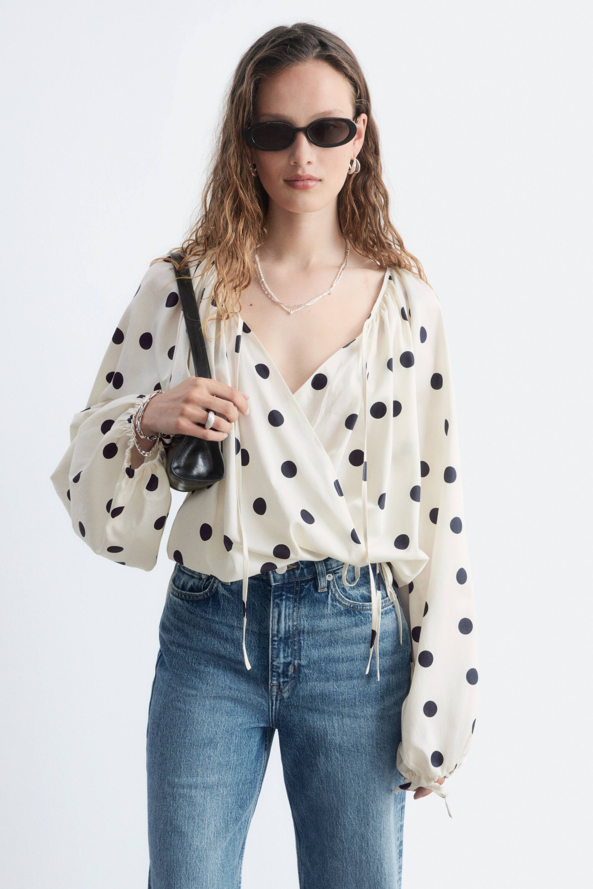 Größeres Bild ansehen: Die cremefarbene Bluse ist mit einem schwarzen Polka-Dot-Muster versehen, einem V-Ausschnitt mit Kordelzug-Detail und langen, voluminösen Ärmeln mit elastischen Bündchen. Sie ist mit einer Wickeloptik vorne und einem verstellbaren Kordelzug am Saum gestaltet und wird in blaue Jeans gesteckt getragen.
