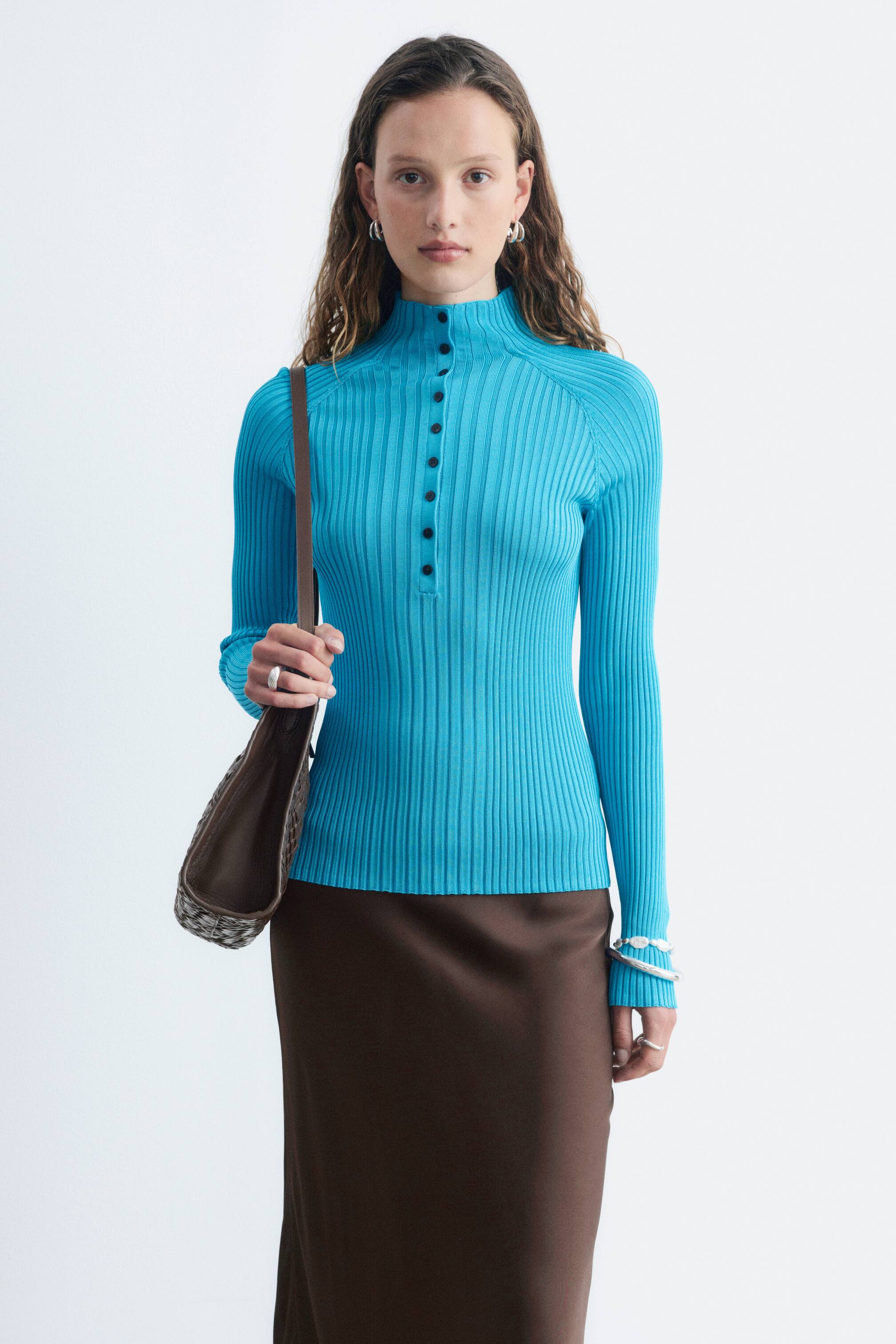 View larger image: A bright blue, slim-fit, rib-knitted top, featuring a mock neck and a buttoned half-placket with black buttons, is being worn by the model and styled with a dark brown skirt.