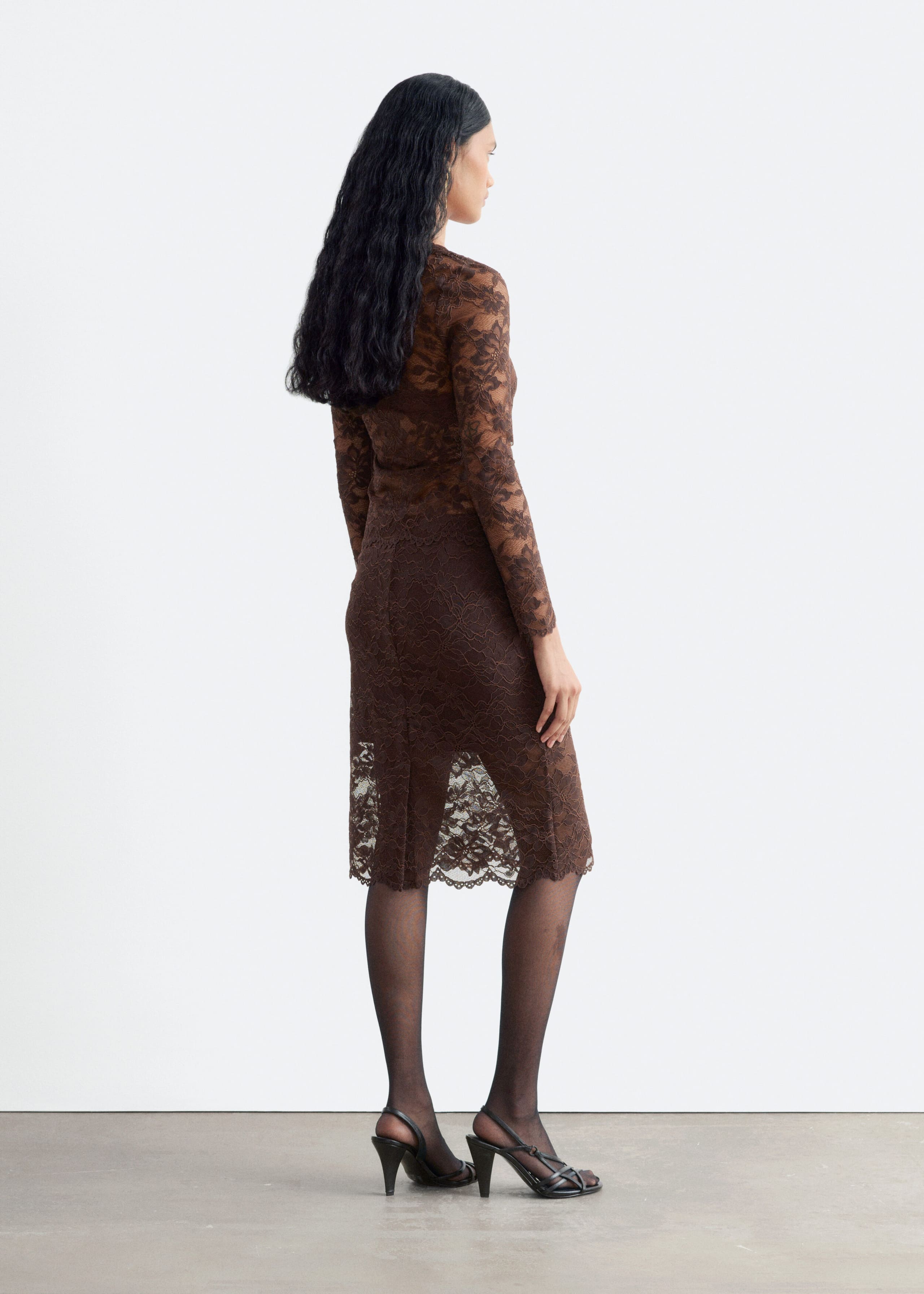 A brown lace skirt featuring a slim, knee-length silhouette and a sheer scalloped hem is being worn by the model. It is styled with a matching brown long-sleeved lace top, sheer black tights, and black high-heeled sandals.
