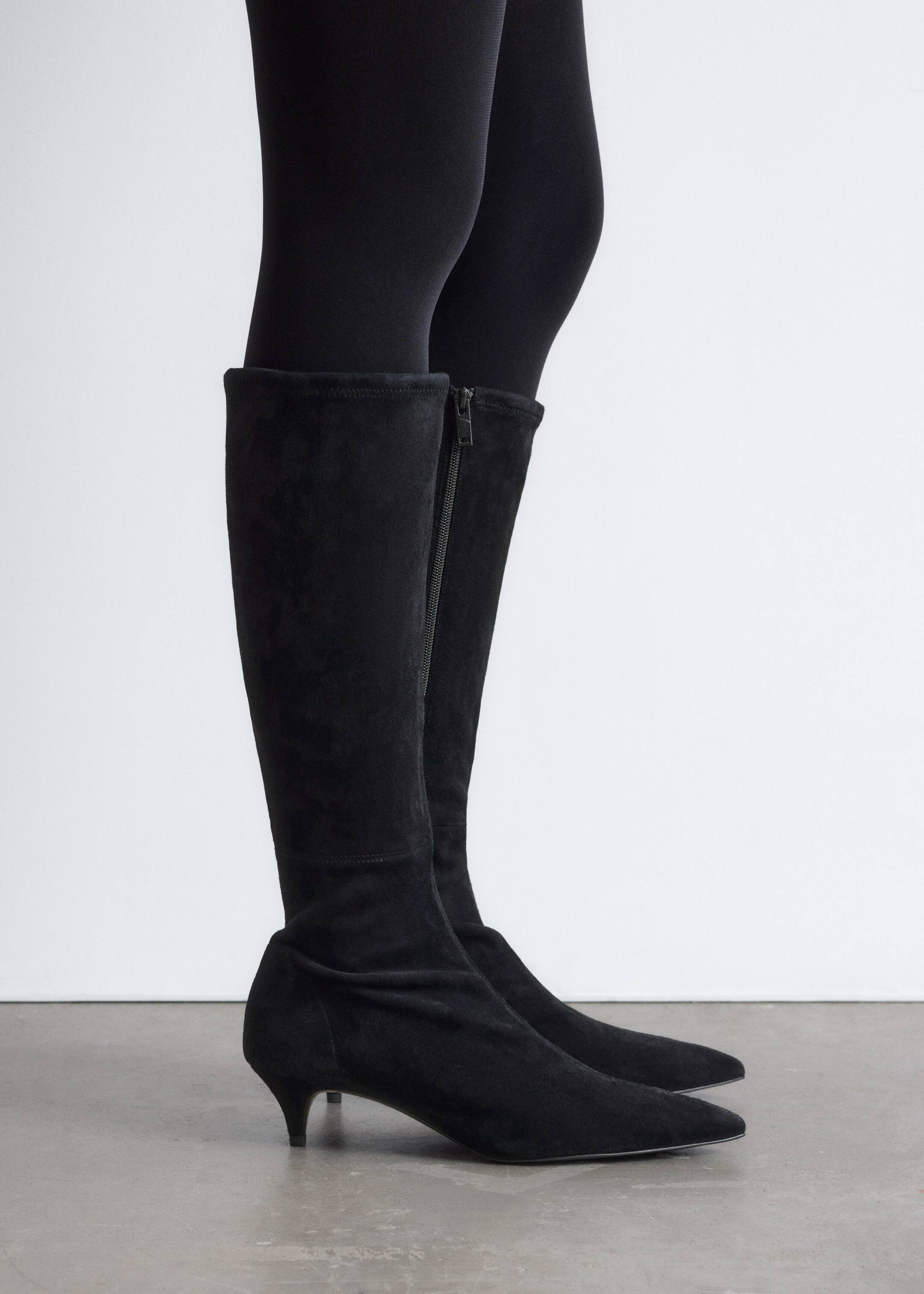 Visa större bild: Svarta knähöga stövlar visas i en sidoprofil, bärs över svarta leggings. Stövlarna är tillverkade av ett svart mockaliknande material, med en spetsig tå och en låg klack. En sidodragkedja löper längs den inre vaden.
