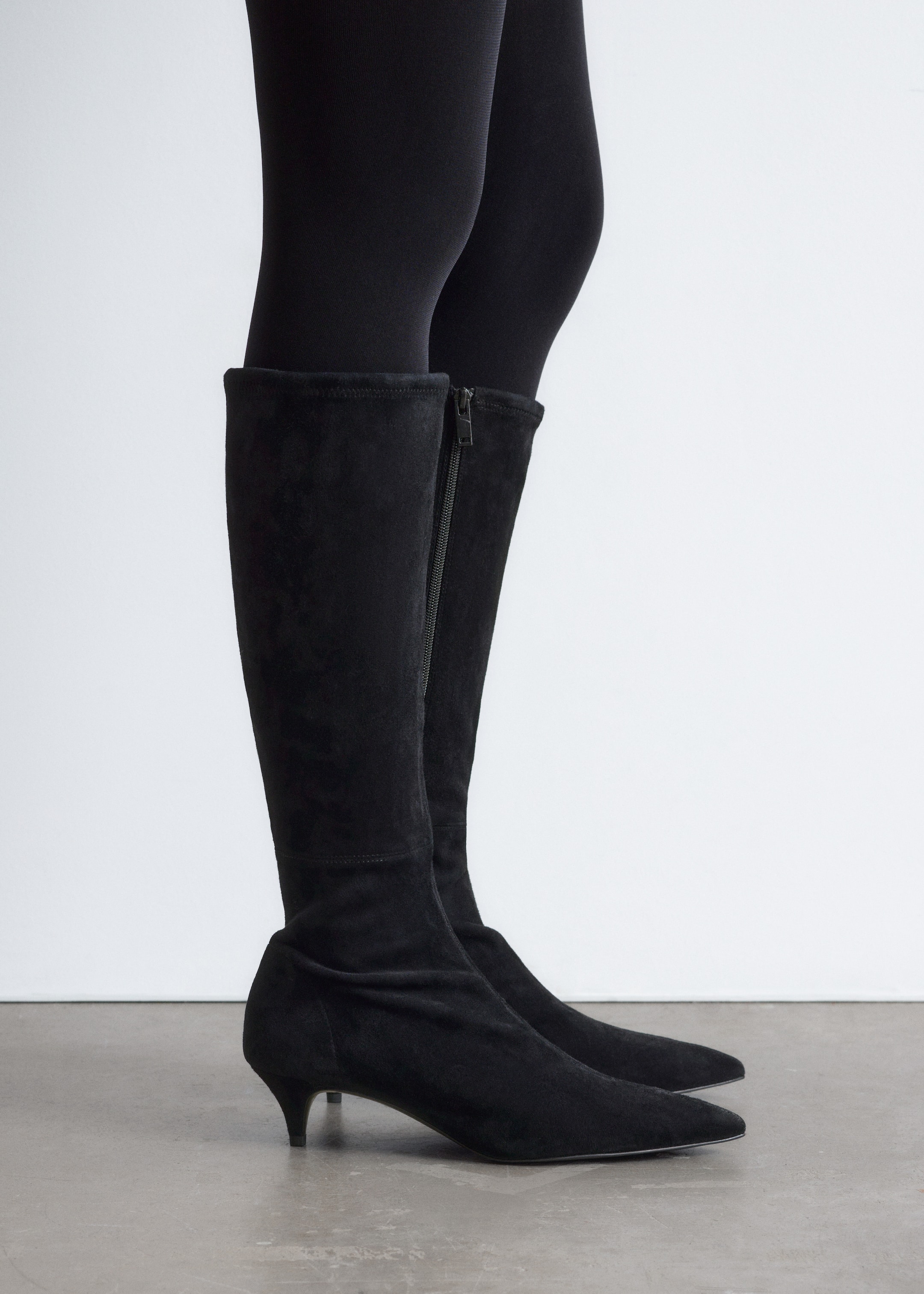 Visa större bild: Svarta knähöga stövlar visas i sidoprofil, bärs över svarta leggings. Stövlarna är tillverkade av ett svart mockaliknande material, med en spetsig tå och en låg klack. En sidodragkedja löper längs den inre vaden.