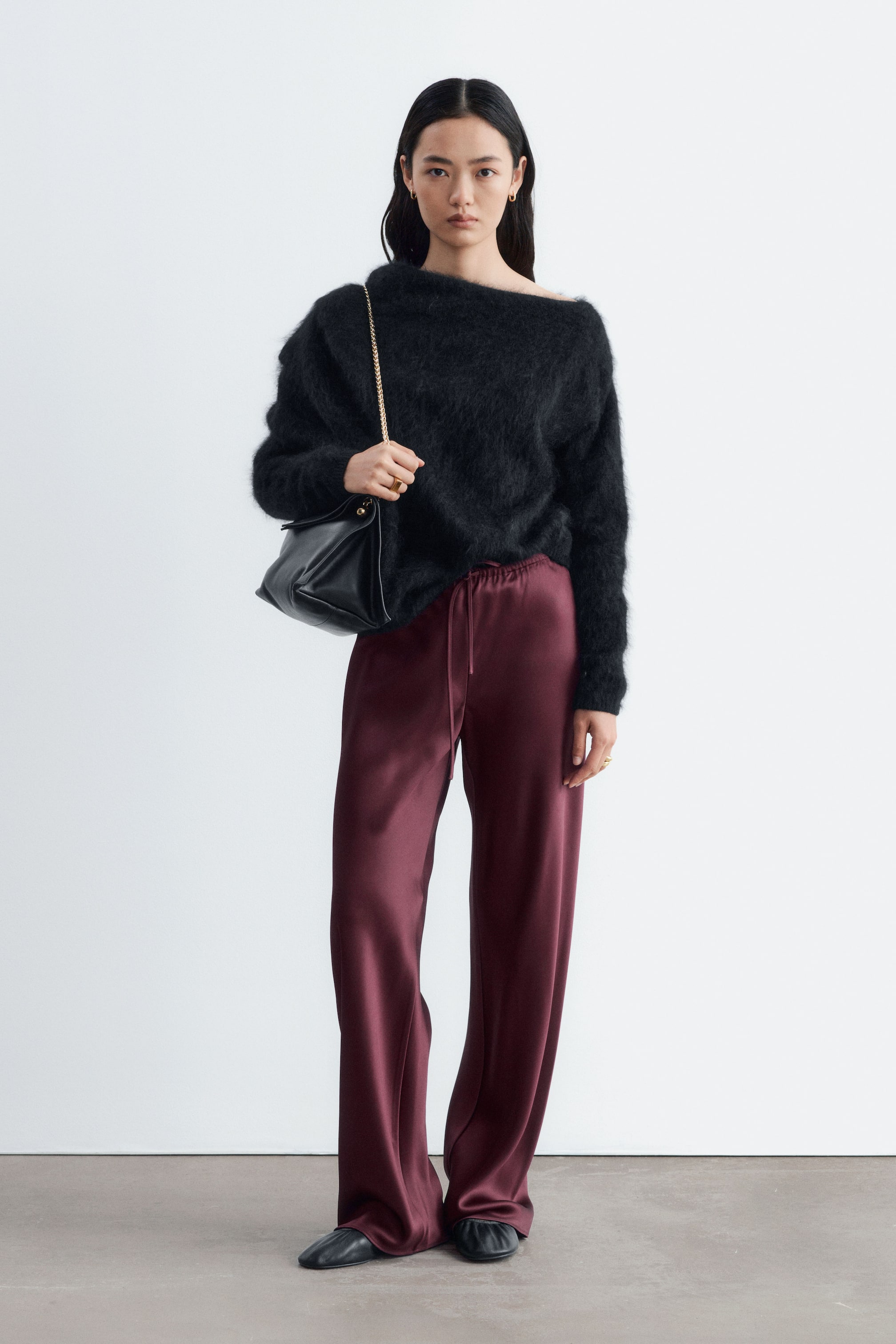 View larger image: The model is wearing burgundy satin trousers featuring a relaxed fit and an elastic drawstring waist. The fluid, wide-leg trousers are styled with a black fuzzy off-the-shoulder jumper and a black leather shoulder bag.