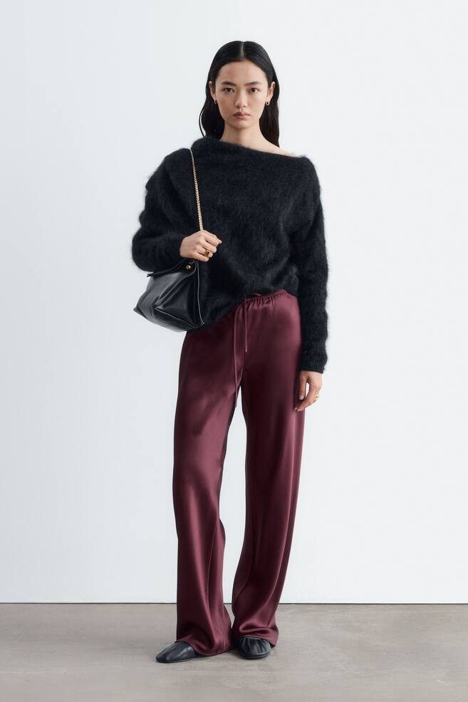 The model is wearing burgundy satin trousers featuring a relaxed fit and an elastic drawstring waist. The fluid, wide-leg trousers are styled with a black fuzzy off-the-shoulder jumper and a black leather shoulder bag. - 1