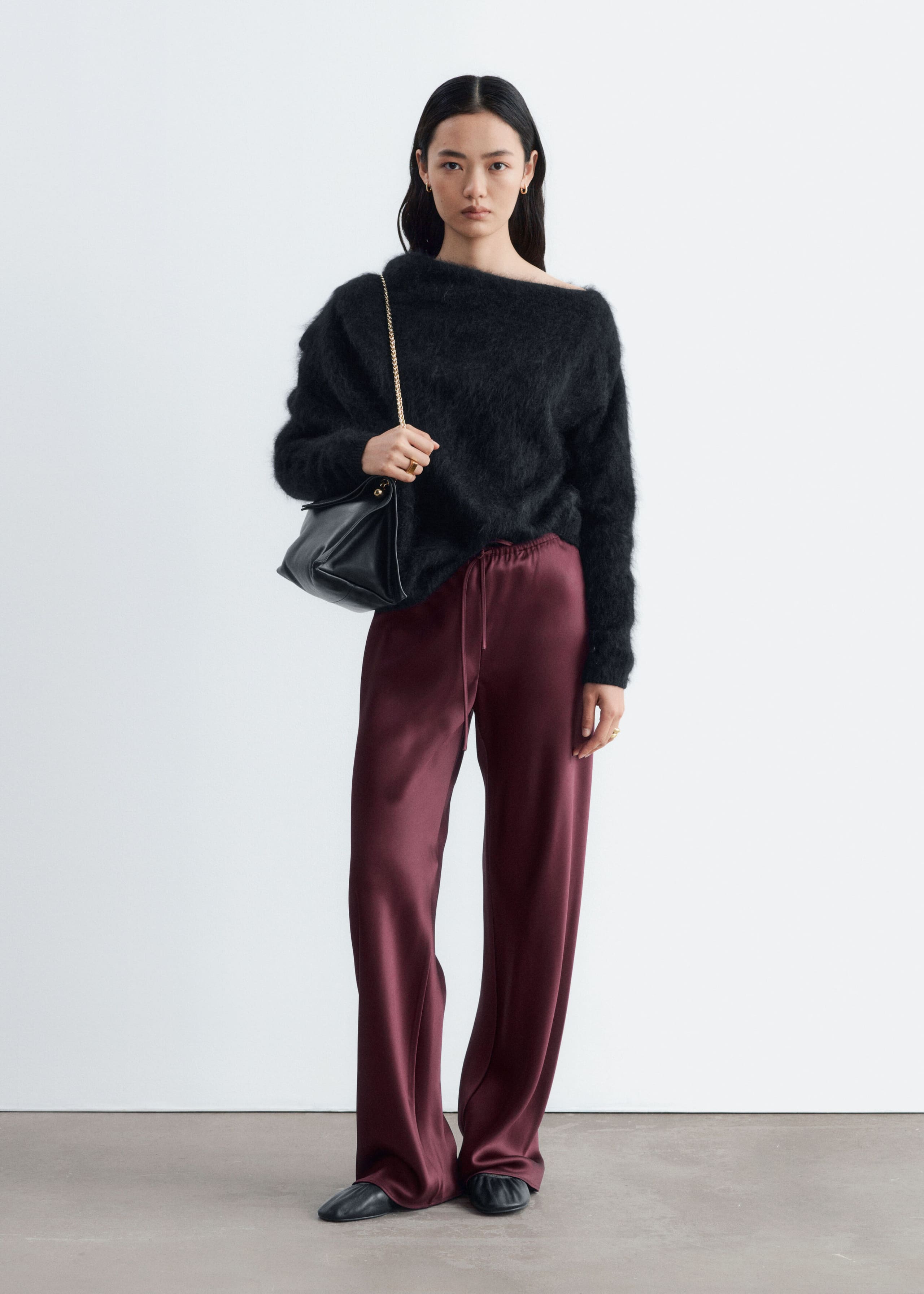 The model is wearing burgundy satin trousers with a relaxed fit and an elastic drawstring waist. The fluid, wide-leg trousers are styled with a black fuzzy off-the-shoulder sweater and a black leather shoulder bag.