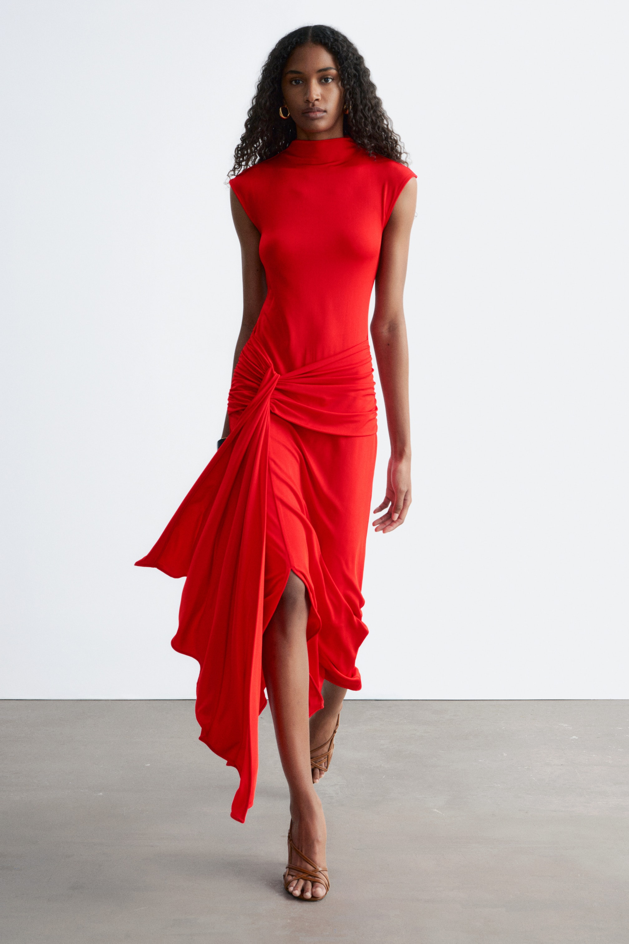 Vis større bilde: En rød kjole med høy hals og kappeermer, med intrikat rynking samlet ved venstre hofte. Fra en svak frontvendt vinkel, flyter det asymmetriske skjørtet dynamisk, og avslører en høy splitt og høyre ben.