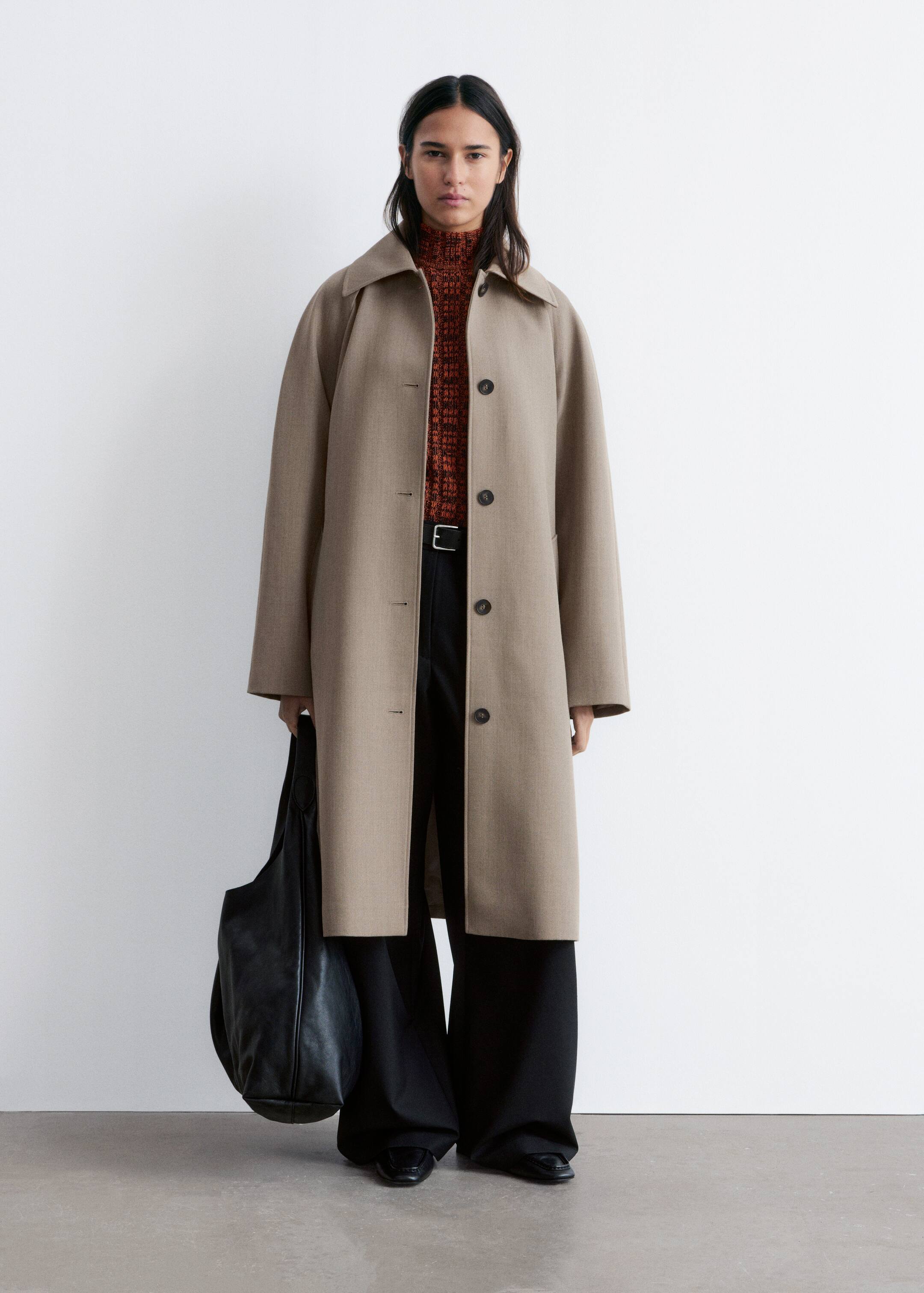 Größeres Bild anzeigen: Der beige lange Mantel zeigt eine legere, gerade Silhouette und wird in frontaler Ansicht präsentiert. Er ist mit einem spitzen Kragen und einer vorderen Vier-Knopf-Leiste mit dunklen Knöpfen versehen und wird derzeit offen über einem gemusterten orangefarbenen Rollkragenpullover und einer schwarzen Hose getragen.