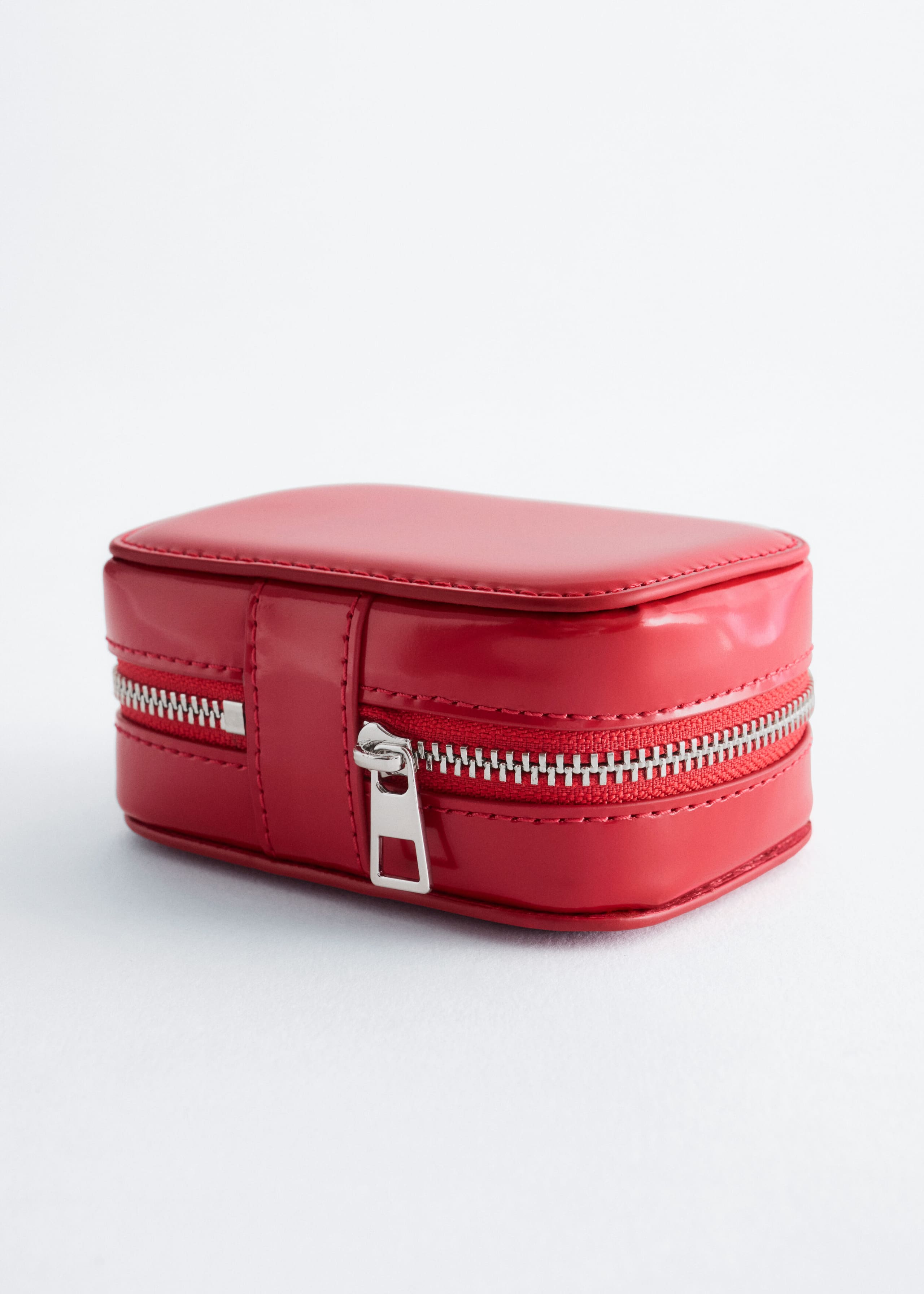 A small, structured red case is displaying a rectangular shape with rounded corners, a smooth glossy finish, and tonal topstitching. A silver-toned metal zip is running horizontally around the centre, featuring a rectangular zip pull on a vertical strap detail.