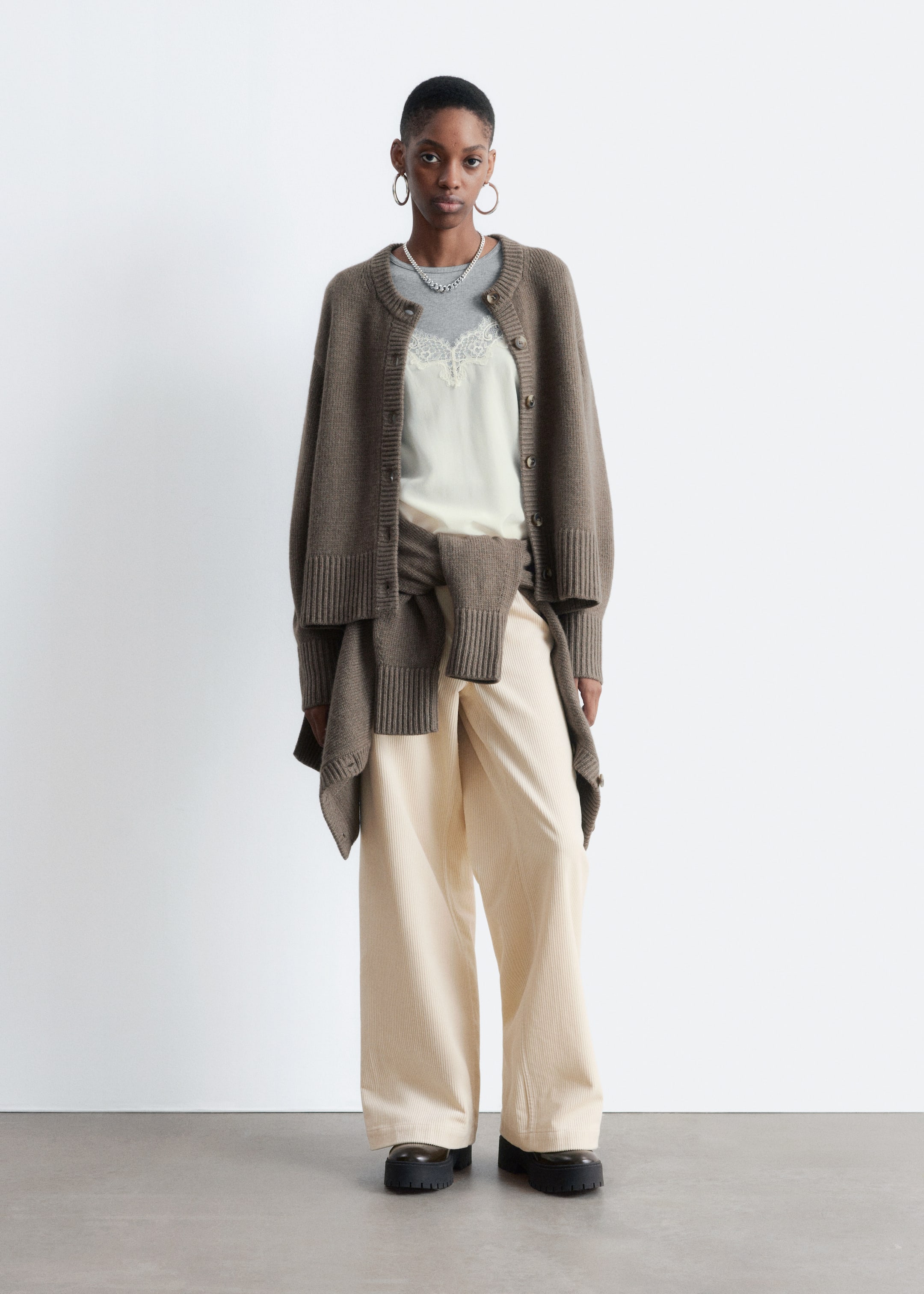 Visualizza immagine più grande: Un cardigan marrone viene presentato da un'angolazione frontale, con un cardigan indossato aperto, caratterizzato da uno scollo rotondo, una chiusura a bottoni frontale e polsini e orlo a coste. Un secondo cardigan marrone abbinato viene anche legato intorno alla vita, completando la t-shirt grigia a strati, il top crema con rifiniture in pizzo, i pantaloni a coste crema a gamba larga e le scarpe nere massicce.