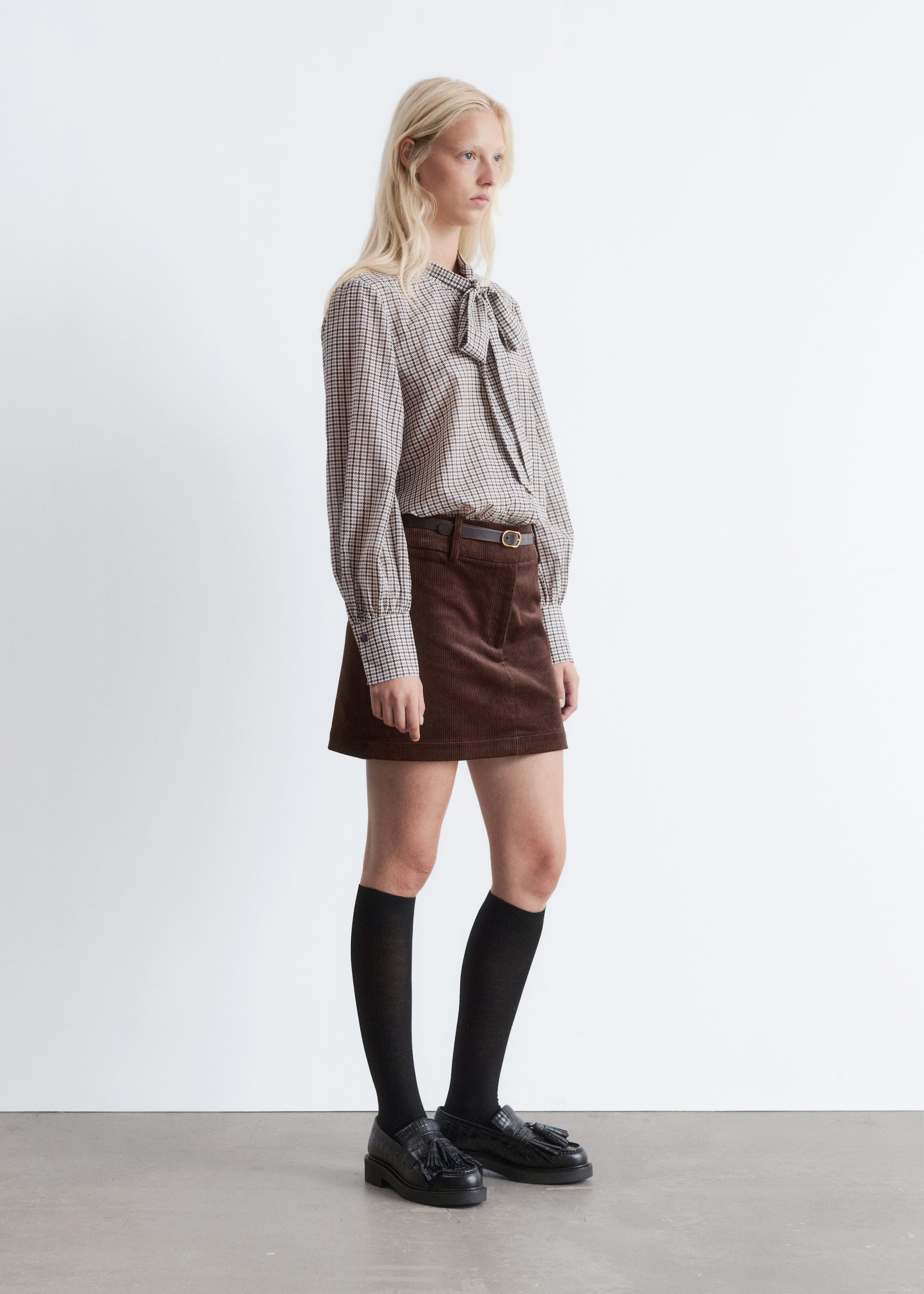 Grösseres Bild ansehen: Eine braun-beige karierte Bluse mit einer Schleifenkragenkrawatte und langen Puffärmeln mit geknöpften Manschetten wird im Seitenprofil getragen, in einen braunen Cord-A-Linien-Minirock gesteckt. Der Rock hat ein dezentes Frontklappendetail und ist an der Taille mit einem dünnen braunen Gürtel gerafft.