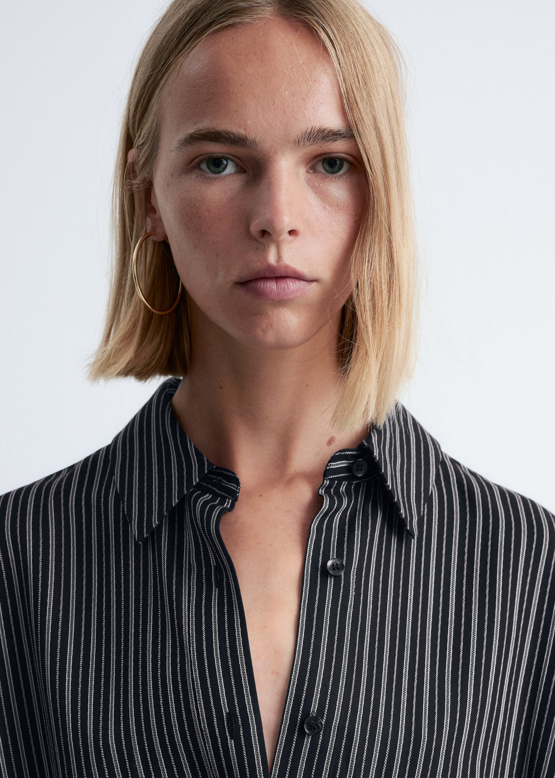 Agrandir l'image: Une chemise noire avec des rayures fines verticales blanches est présentée de face. Elle est dotée d'un col pointu et d'une patte de boutonnage à l'avant, avec les boutons du haut défaits pour créer une ouverture en V.