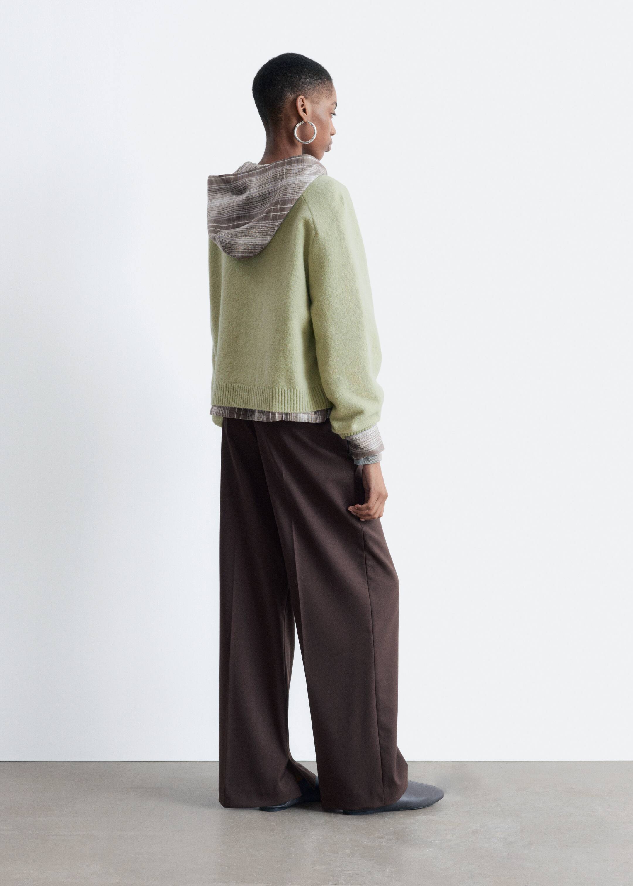 View larger image: A light green hooded sweater, appearing fuzzy, is being layered over a brown and beige plaid shirt, with the plaid visible at the hood, cuffs, and hem. It is being paired with dark brown, wide-leg trousers, all shown from the back right side.