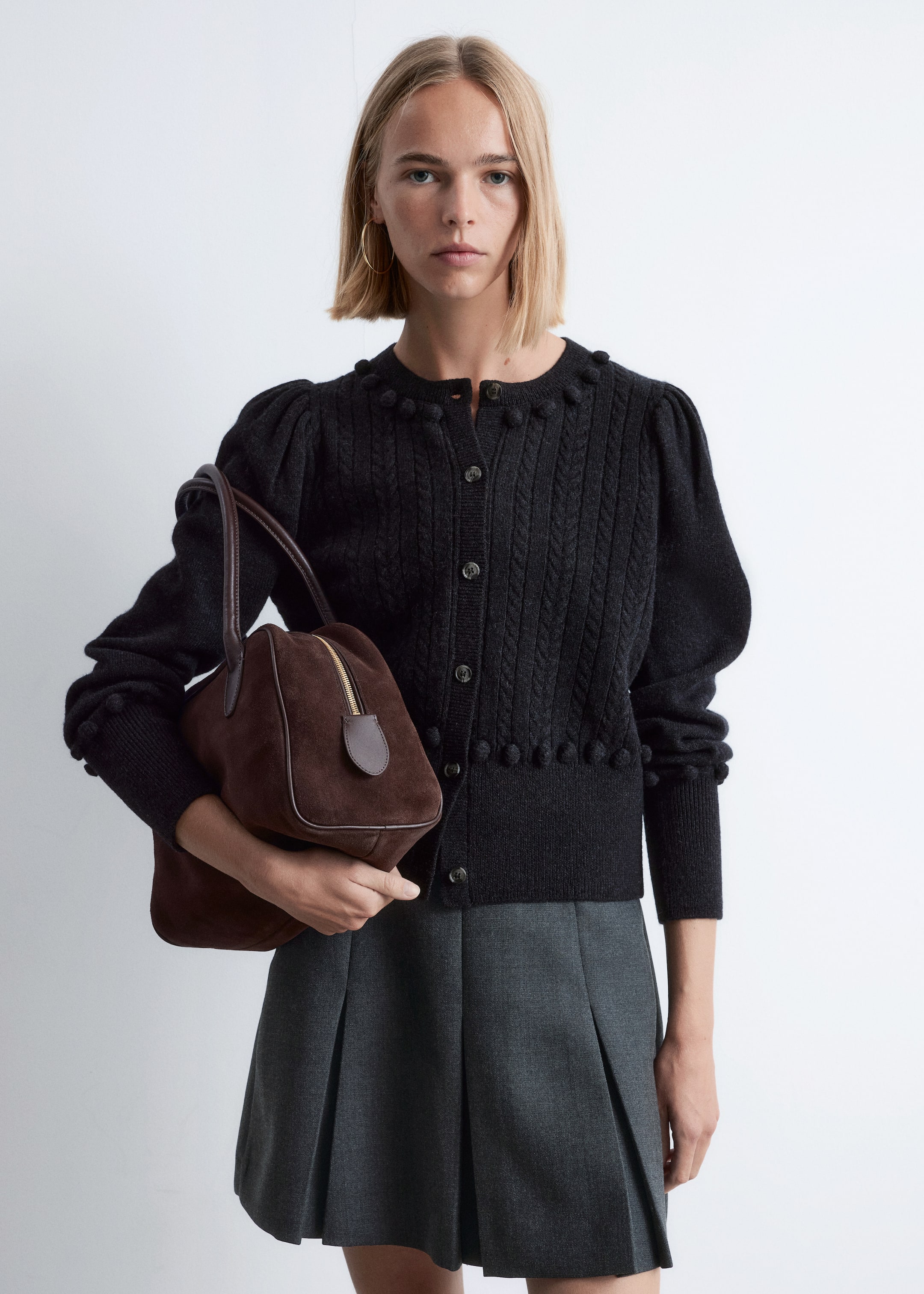 View larger image: A black cable knit cardigan with a round neckline, puffed sleeves, and pom-pom detailing along the neckline, hem, and cuffs is being presented from a front view. It is being styled with a dark gray pleated mini skirt, and a brown suede structured bag with a gold-toned top zipper and two handles is being held.