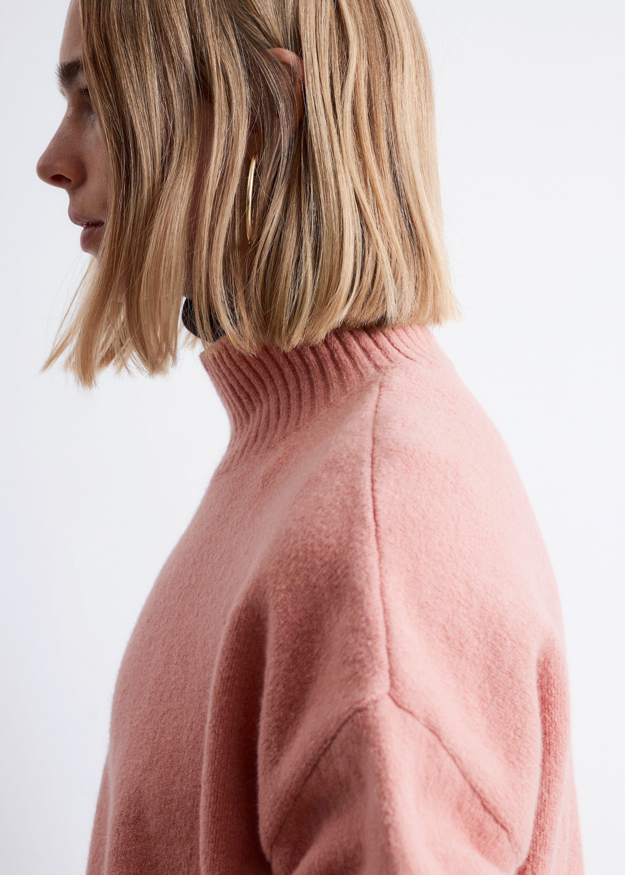 Größeres Bild anzeigen: Ein rosa Pullover wird aus einer Profilansicht gezeigt, wobei seine weiche, flauschige Strickstruktur und die entspannte Passform mit einer sichtbaren Schulternaht hervorgehoben werden. Er verfügt über einen gerippten Stehkragen, unter dem eine dunkle Schicht sichtbar ist.