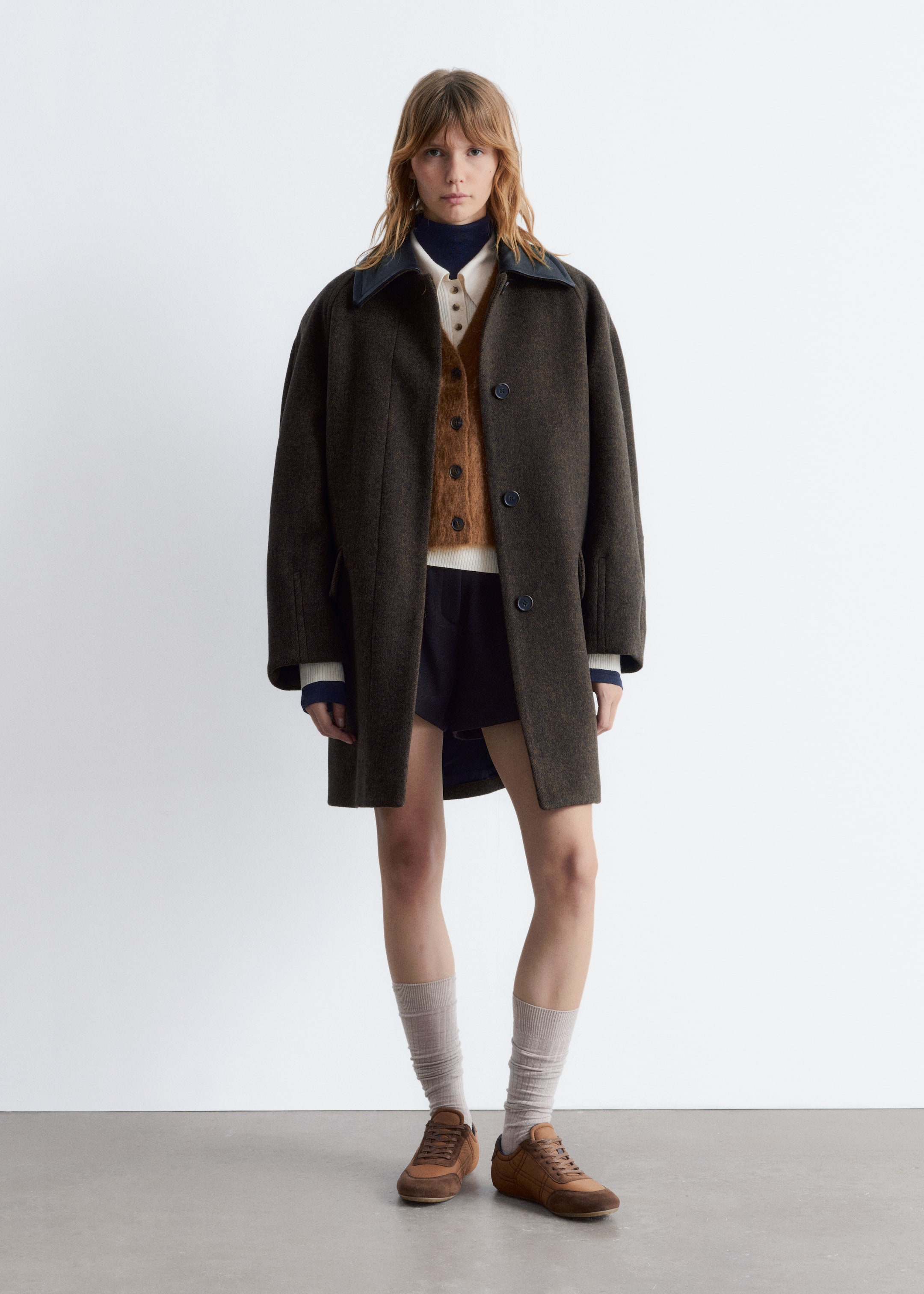 Grotere afbeelding bekijken: Een donker olijfgroene oversized jas wordt gepresenteerd, naar voren gericht, met een donker contrasterende spreidkraag, verlaagde schouders en donkere ronde knopen aan de voorkant. Deze mid-thigh-lange jas wordt over een wit shirt, een donkerblauwe coltrui en een bruin vest gedragen.