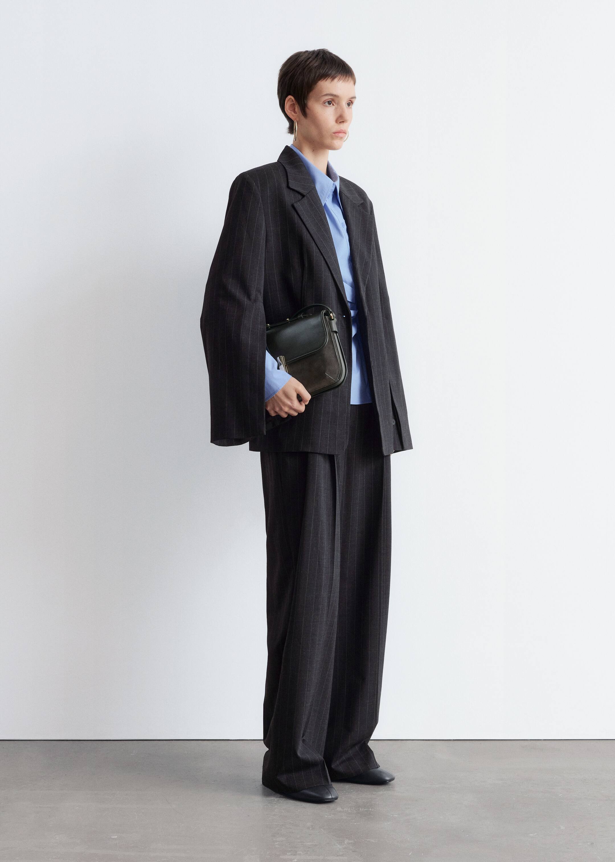 Größeres Bild ansehen: Eine Person steht in einem leichten rechten Winkel und trägt einen dunklen Nadelstreifenanzug, der aus einem übergroßen Blazer mit Kerbrevers und weit geschnittenen Hosen besteht. Unter dem Blazer ist ein hellblaues Hemd mit Kragen sichtbar, und sie halten eine dunkle Lederschultertasche mit Klappenverschluss und goldfarbener Schnalle.