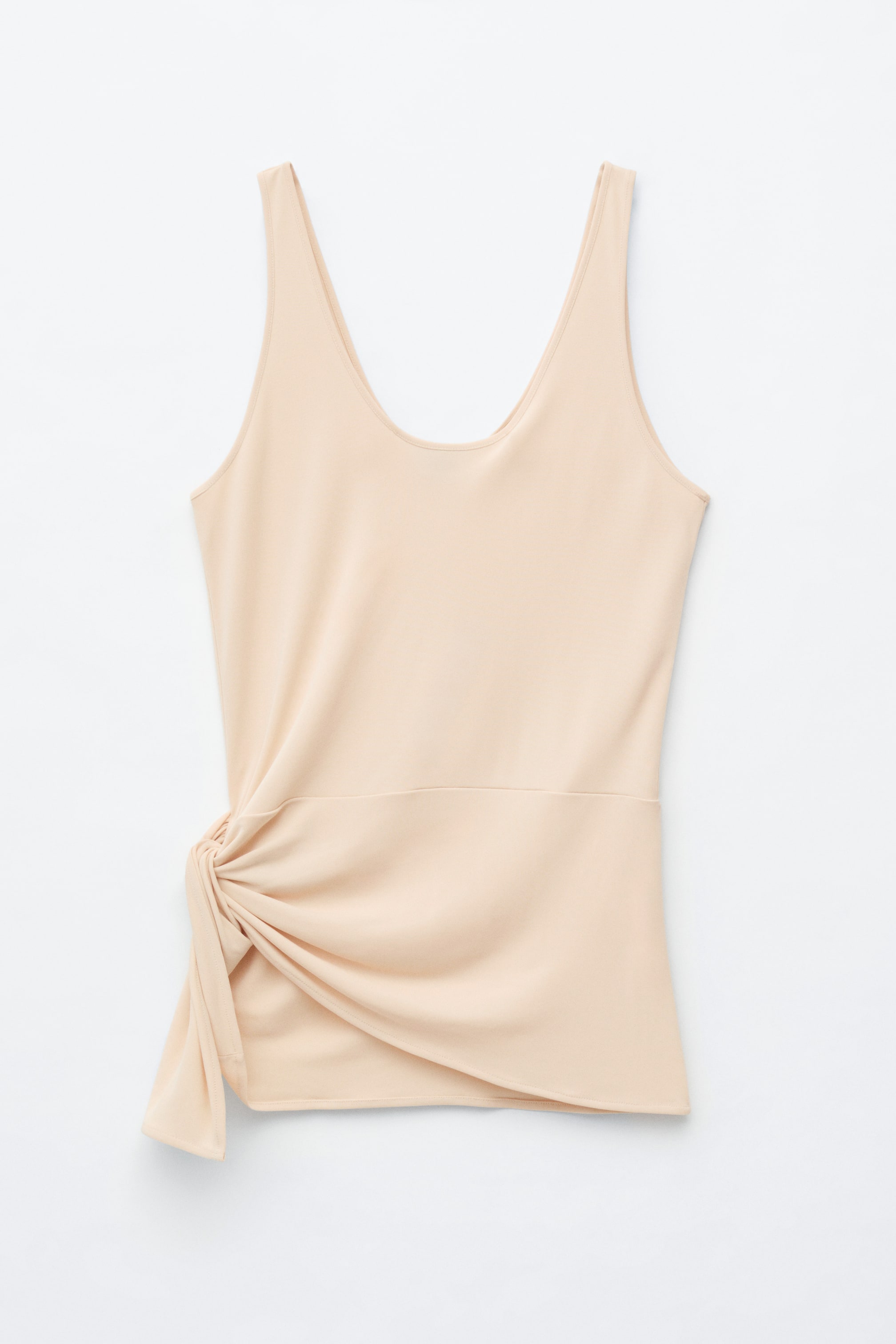 Vis større bilde: En lys beige tanktopp viser sin front, med en rund halsutringning og brede stropper. Venstre side av falden samler seg i en knute, som skaper draperinger og en asymmetrisk avslutning.