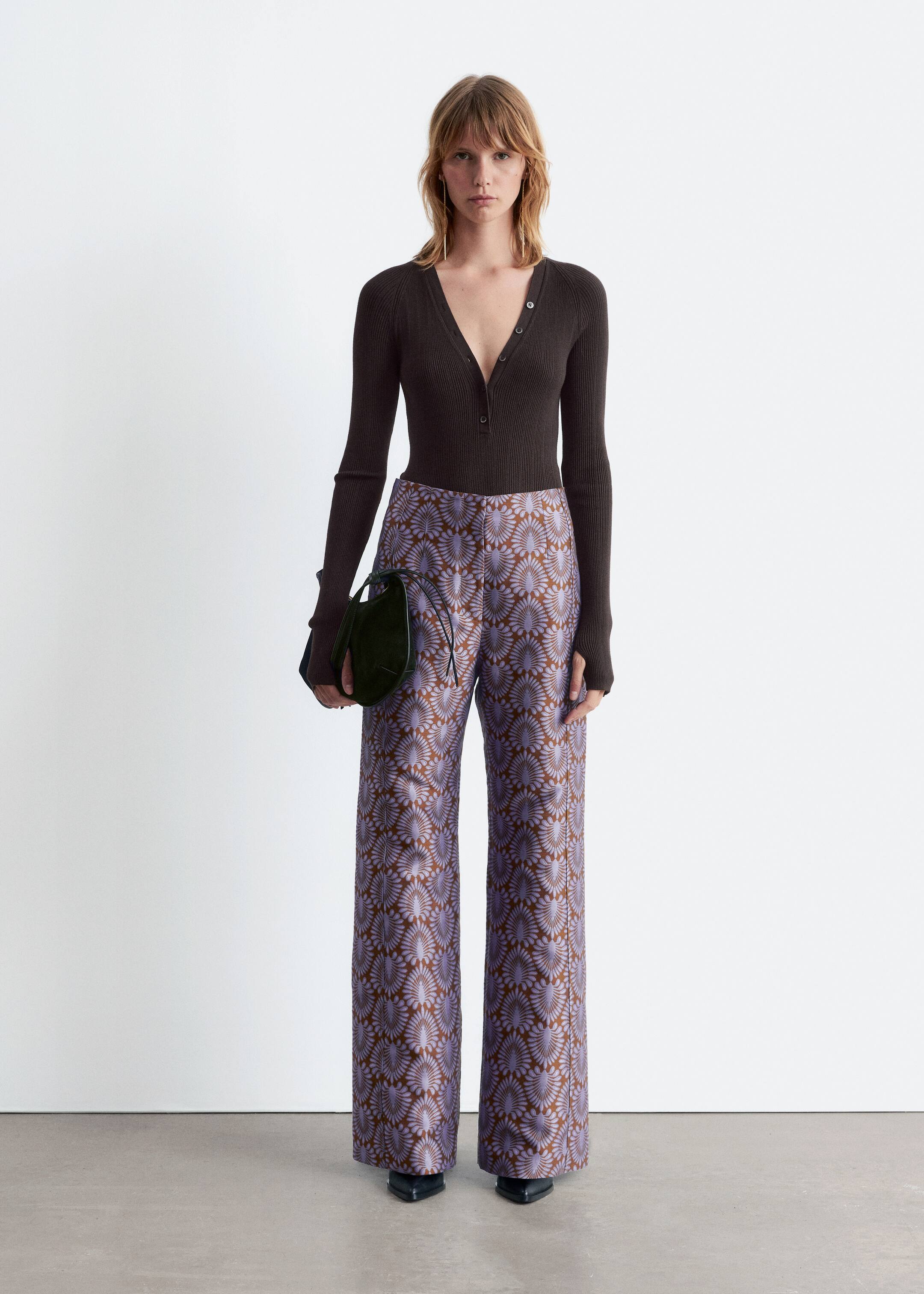 View larger image: A dark brown, slim-fit rib-knit bodysuit featuring a V-neckline with buttons and long raglan sleeves with thumbhole openings is being worn by the model, styled tucked into patterned wide-leg pants.