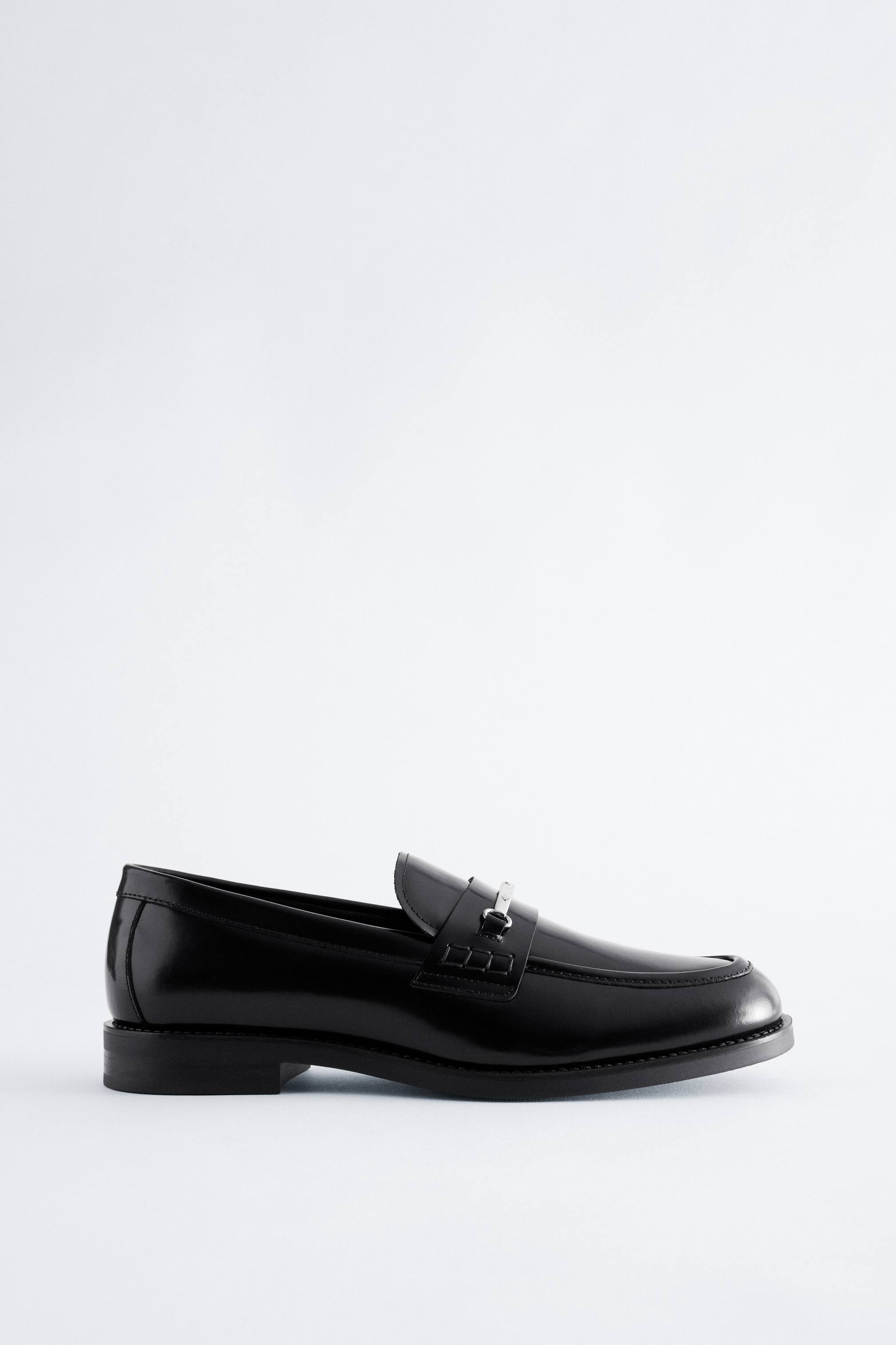 Ver imagem maior: Um mocassim preto em pele polida está virado para a direita, em vista de perfil lateral, exibindo um bico amendoado, um salto baixo em bloco e um detalhe metálico prateado em forma de horsebit na tira do peito do pé.
