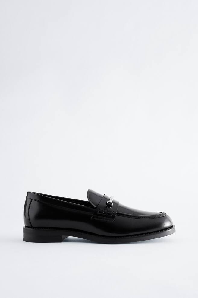 Een zwart gepolijste leren loafer is van opzij naar rechts gericht, met een amandelvormige neus, een lage blokhak en een zilverkleurig metalen horsebit-detail over de wreefband. - 1