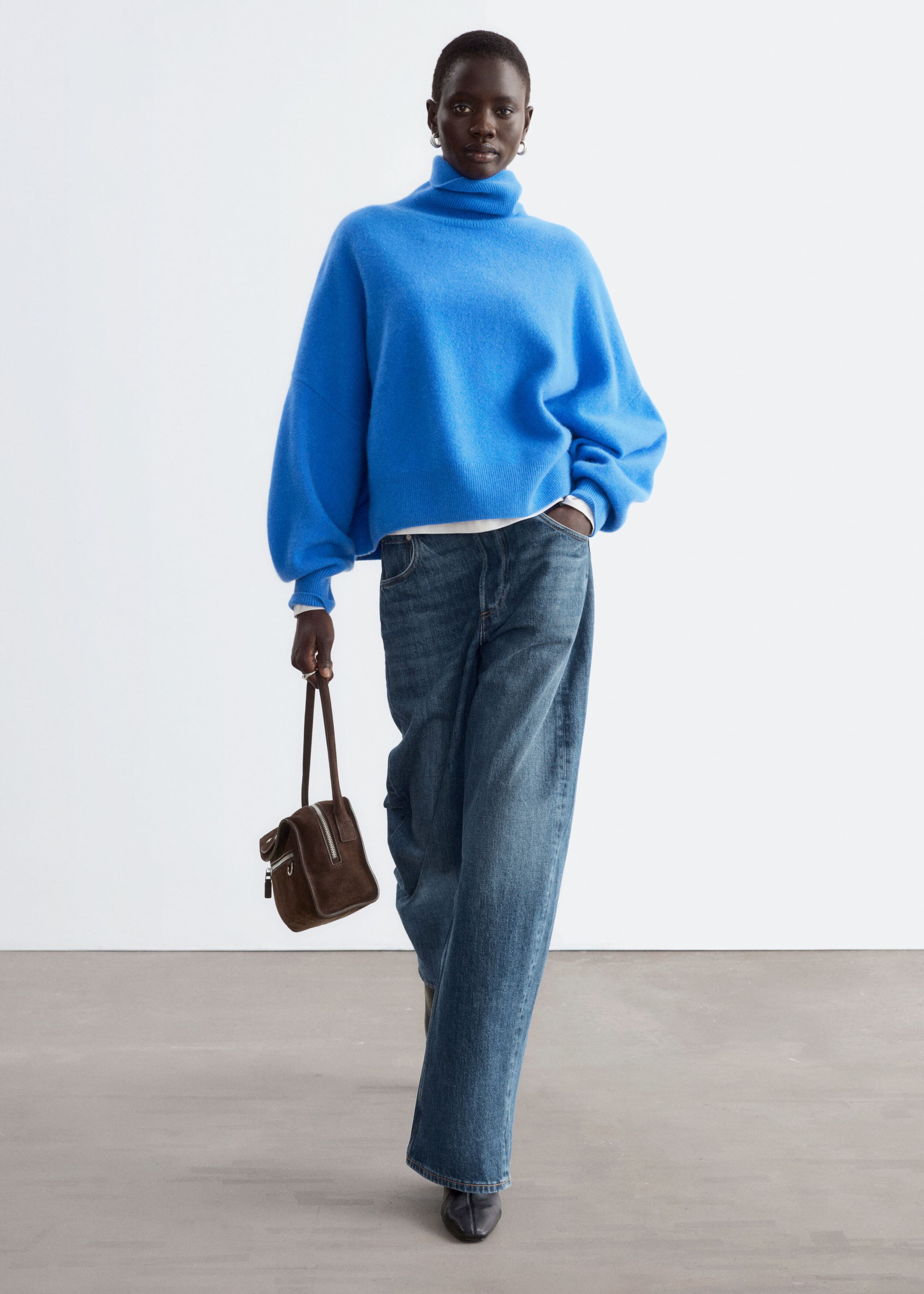 An oversized bright blue jumper featuring a high folded roll neck and dropped shoulders is being worn by the model. The jumper is styled with wide-leg blue jeans and a small brown handbag.