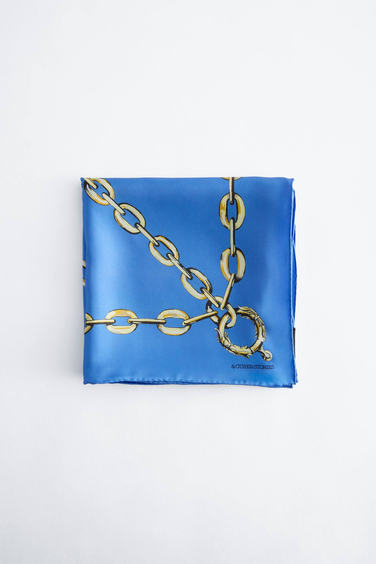 Una sciarpa di seta blu Ã¨ mostrata piegata a forma di quadrato, con vista frontale della sua stampa a catena dorata. Il design presenta catene dorate intrecciate e una chiusura circolare decorativa, con il nome del marchio "& OTHER STORIES" stampato in nero nell'angolo in basso a destra.