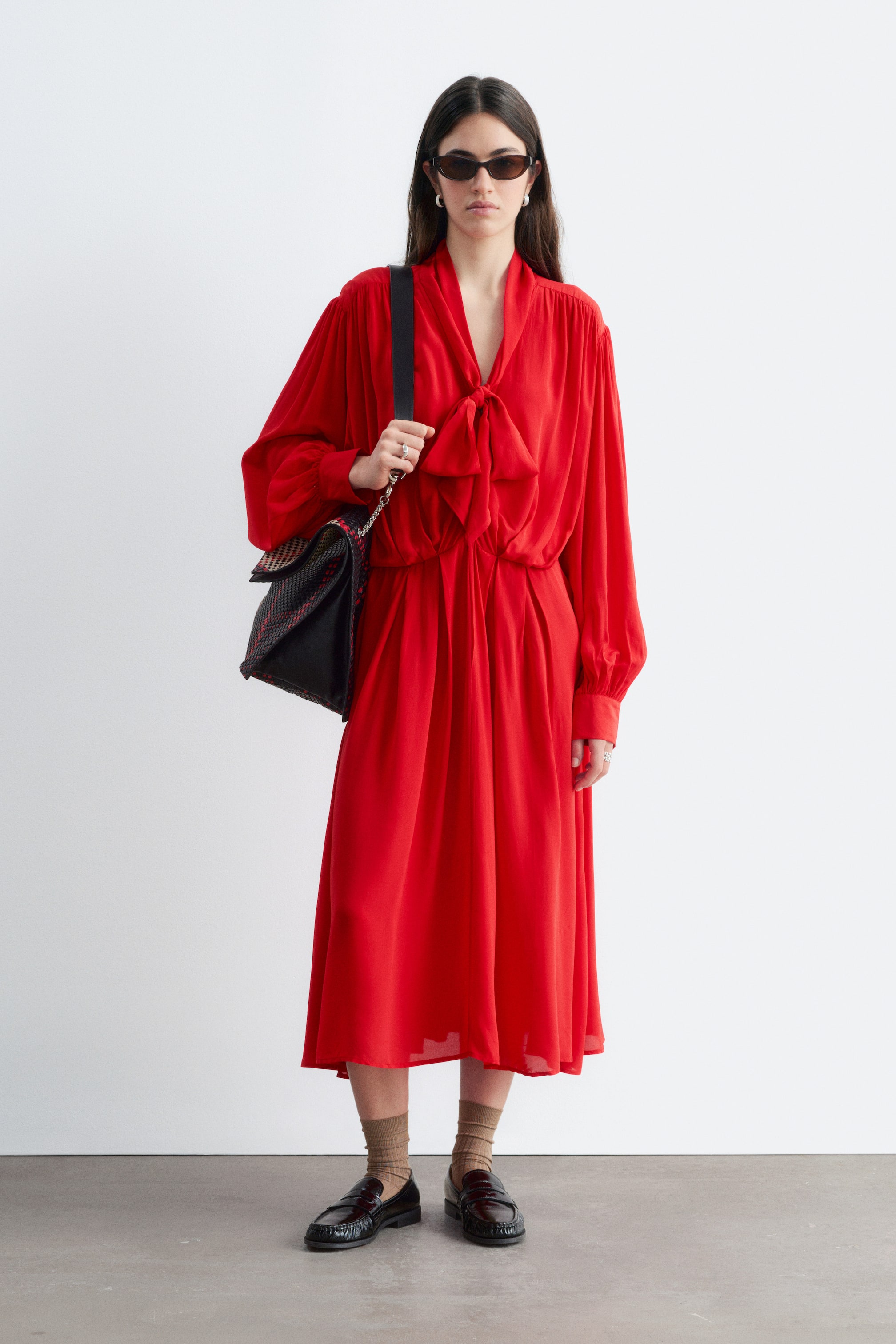 Agrandir l'image: Une robe midi rouge avec un col noué en nœud, des épaules froncées, des manches longues et une taille élastiquée est portée par le mannequin. La robe est associée à un sac bandoulière noir et à des mocassins noirs.