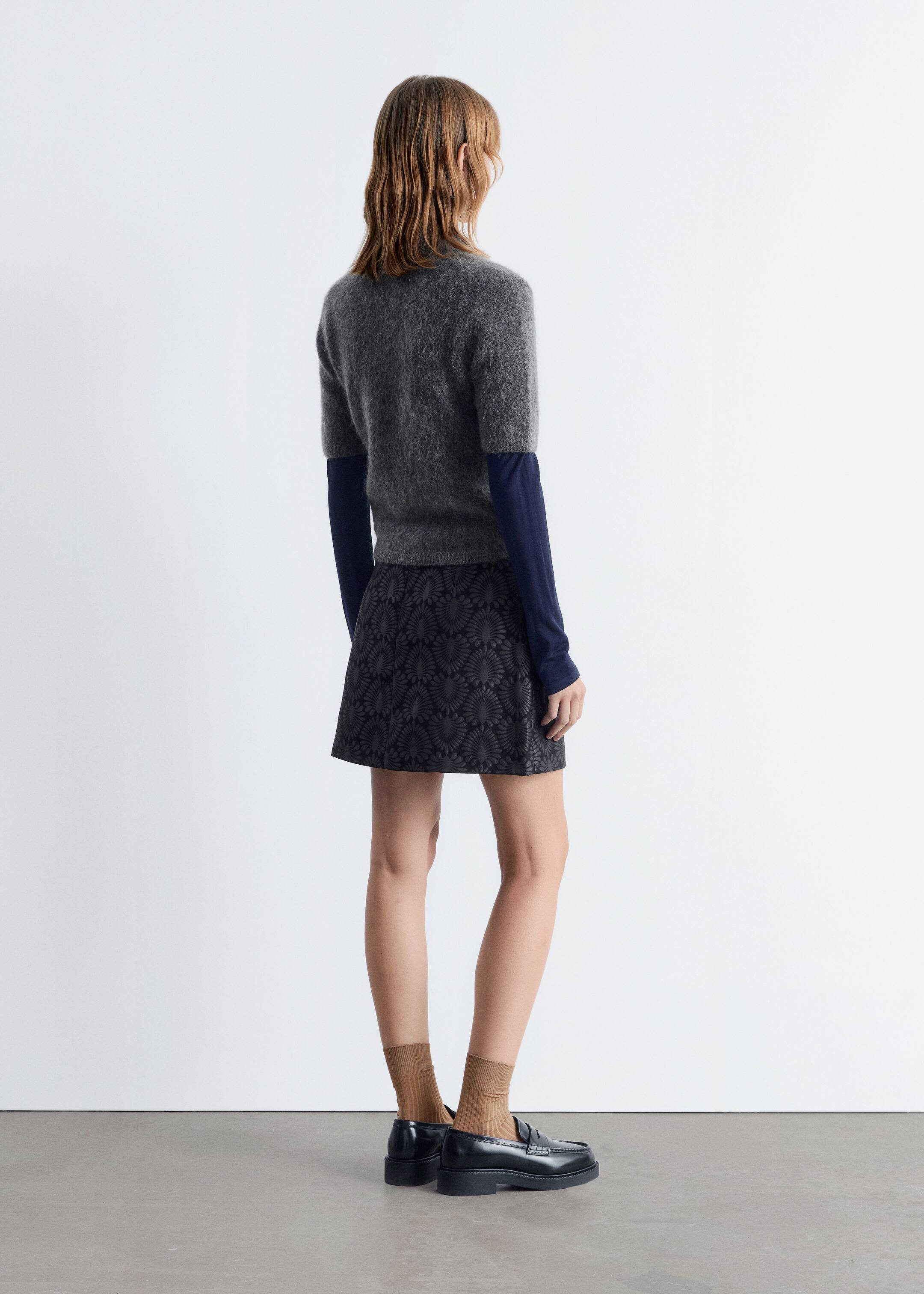 Größeres Bild ansehen: Ein geschichtetes Outfit wird an einer Person präsentiert, die von hinten und leicht nach rechts geneigt betrachtet wird. Es besteht aus einem grauen, kurzärmeligen, flauschigen Pullover, der über einem marineblauen langärmeligen Oberteil getragen wird, kombiniert mit einem dunklen A-Linien-Minirock mit einem subtilen fächerartigen Muster und abgerundet mit schwarzen klobigen Loafern, die mit gerippten braunen Socken getragen werden.