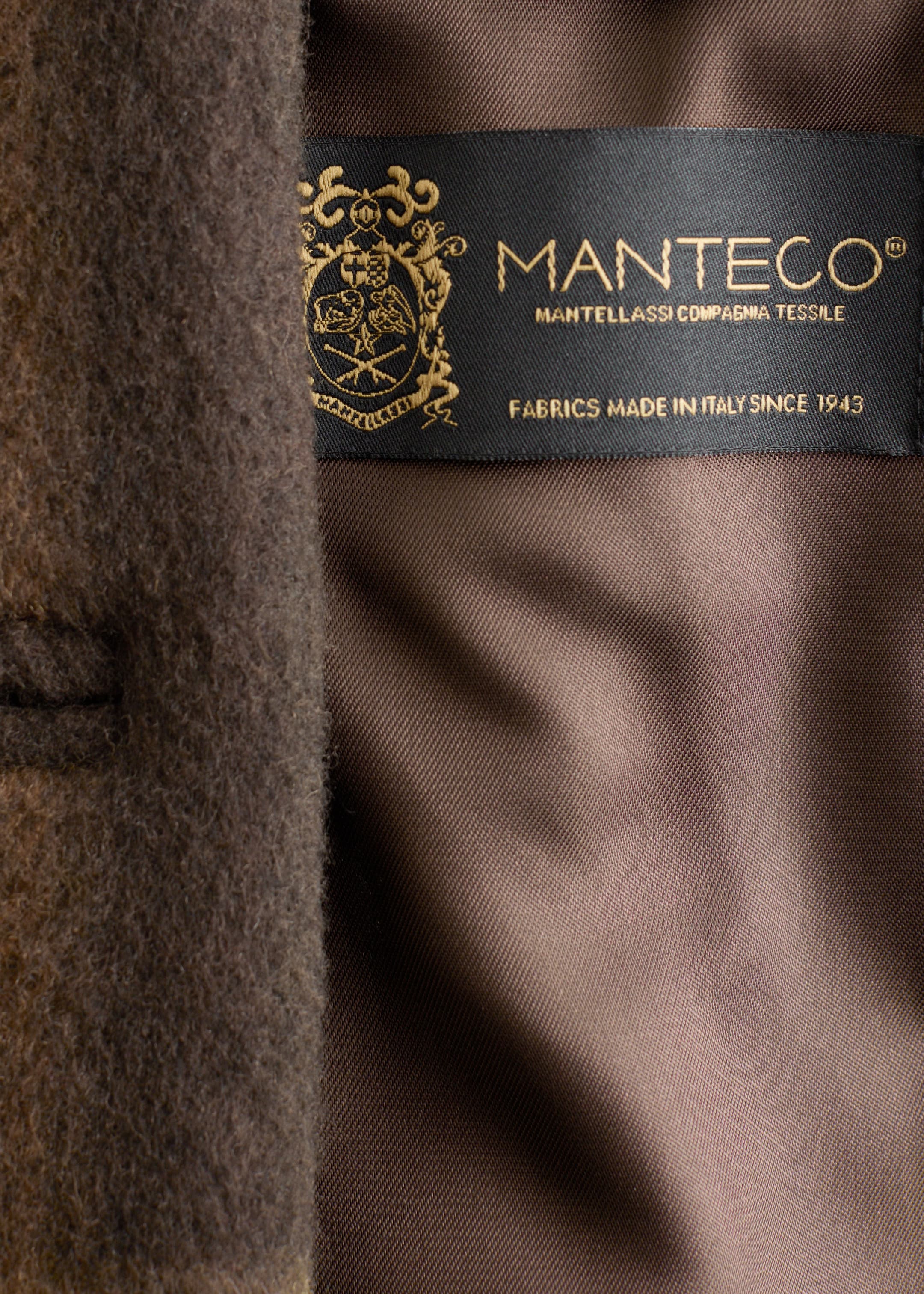 Grotere afbeelding bekijken: Een bruine, donzige buitenstof heeft een zwart geweven label met een gouden embleem en tekst. Het label is genaaid op een gladde, bruine voering en toont "MANTECO" en "FABRICS MADE IN ITALY SINCE 1943."
