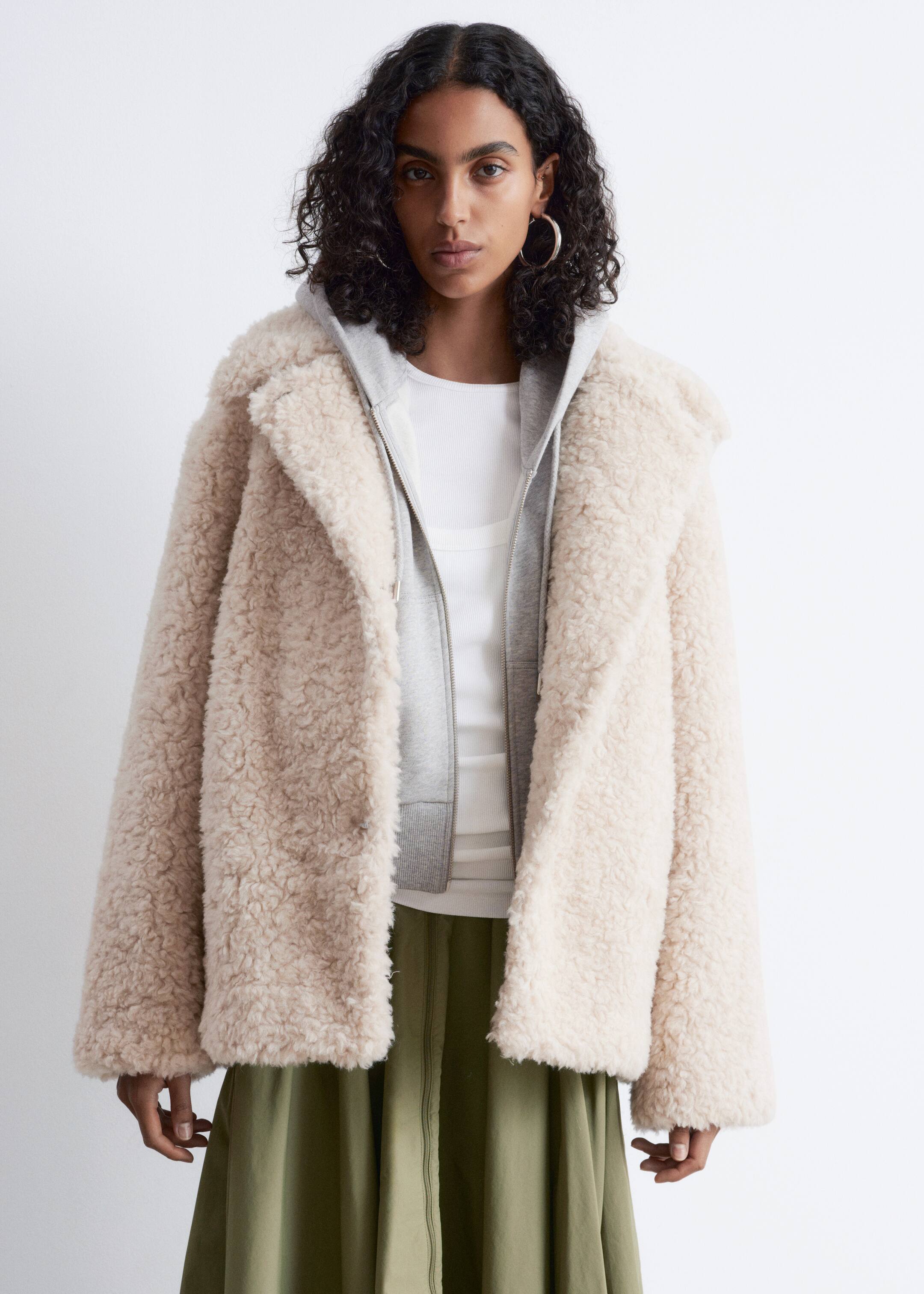 Größeres Bild ansehen: Eine cremefarbene Kunstpelzjacke wird von vorne präsentiert und offen über einem grauen Kapuzenpullover mit sichtbaren gerippten Bündchen getragen. Sie verfügt über breite Einkerbungslappen und weite Ärmel.