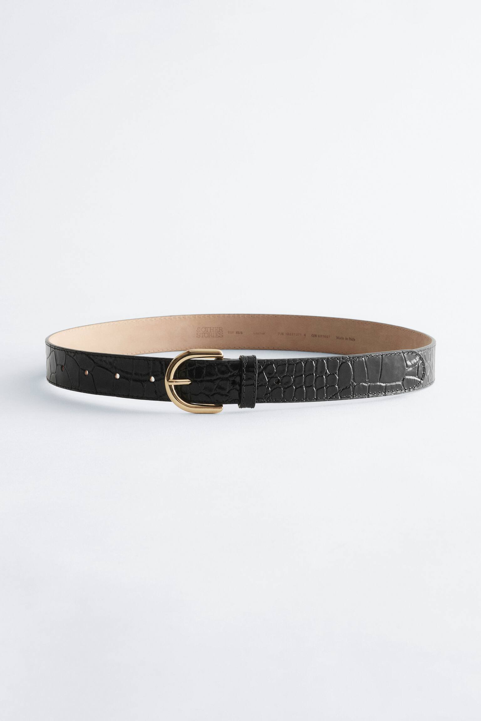 Een zwarte leren riem met crocoprint ligt in een losse cirkel, aan de voorkant zichtbaar met de goudkleurige, afgeronde gesp vastgemaakt. De riem toont aan de buitenzijde een gestructureerd krokodillenpatroon, een bijpassende leren lus en een gladde, tankleurige voering aan de binnenkant.