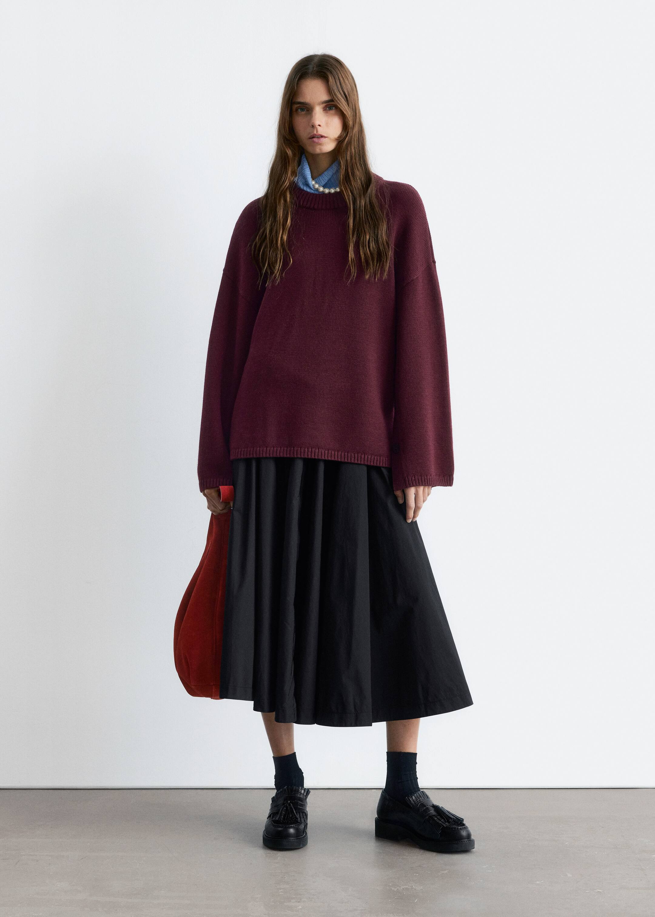 Agrandir l'image: Une tenue complète est présentée, mettant en vedette un pull à col rond bordeaux surdimensionné avec des poignets et un ourlet côtelés, superposé à une chemise à col bleu clair et un collier de perles. Elle est stylée avec une jupe midi plissée noire, des mocassins à glands noirs et un sac rouge non structuré tenu à la main.