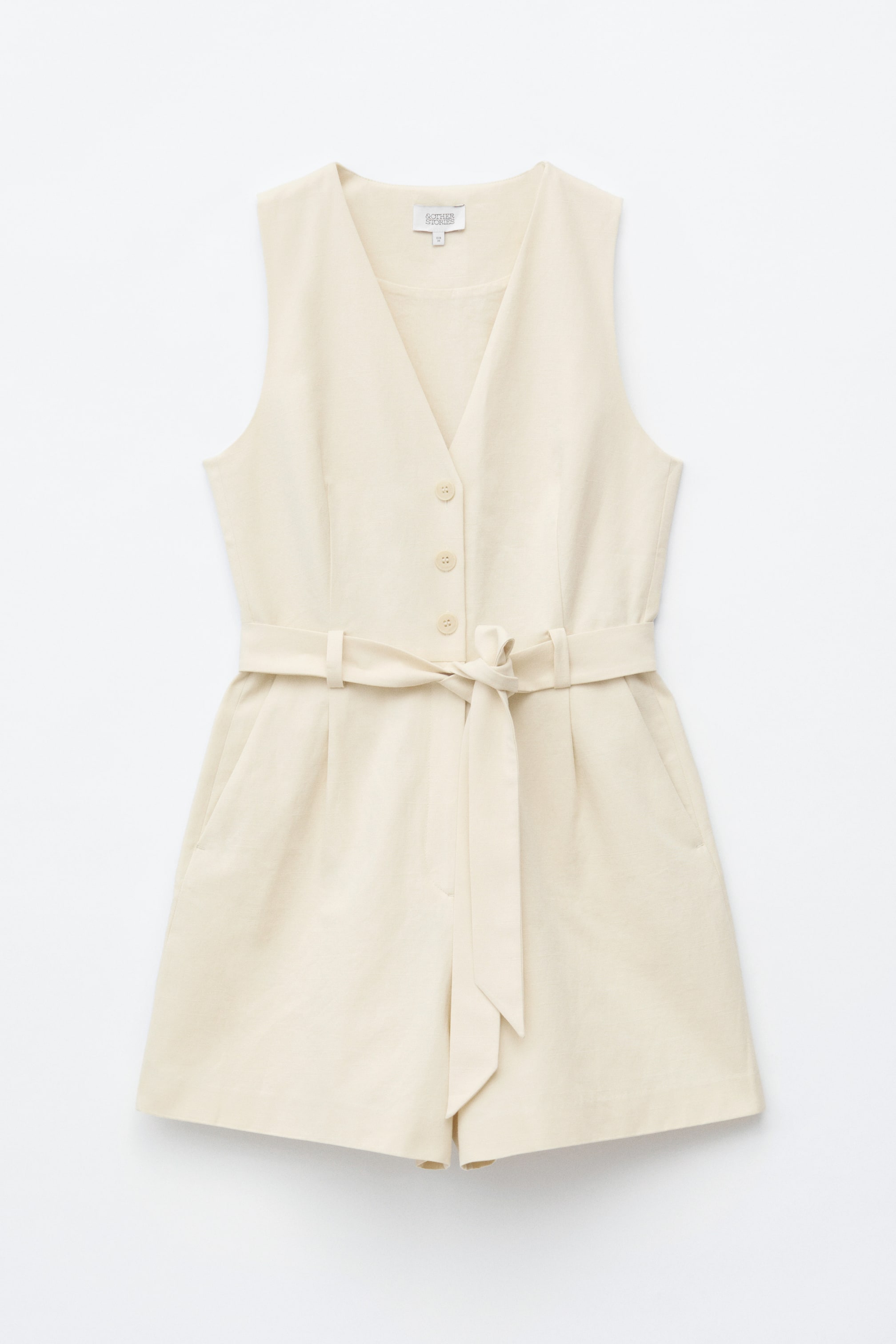 Größeres Bild ansehen: Ein ärmelloser Playsuit in hellem Beige zeigt sich von vorne, mit einem V-Ausschnitt und Knöpfen entlang der Vorderseite. Ein tonaler Stoffgürtel zum Binden betont die Taille, und seitliche Taschen sind an den Shorts sichtbar.