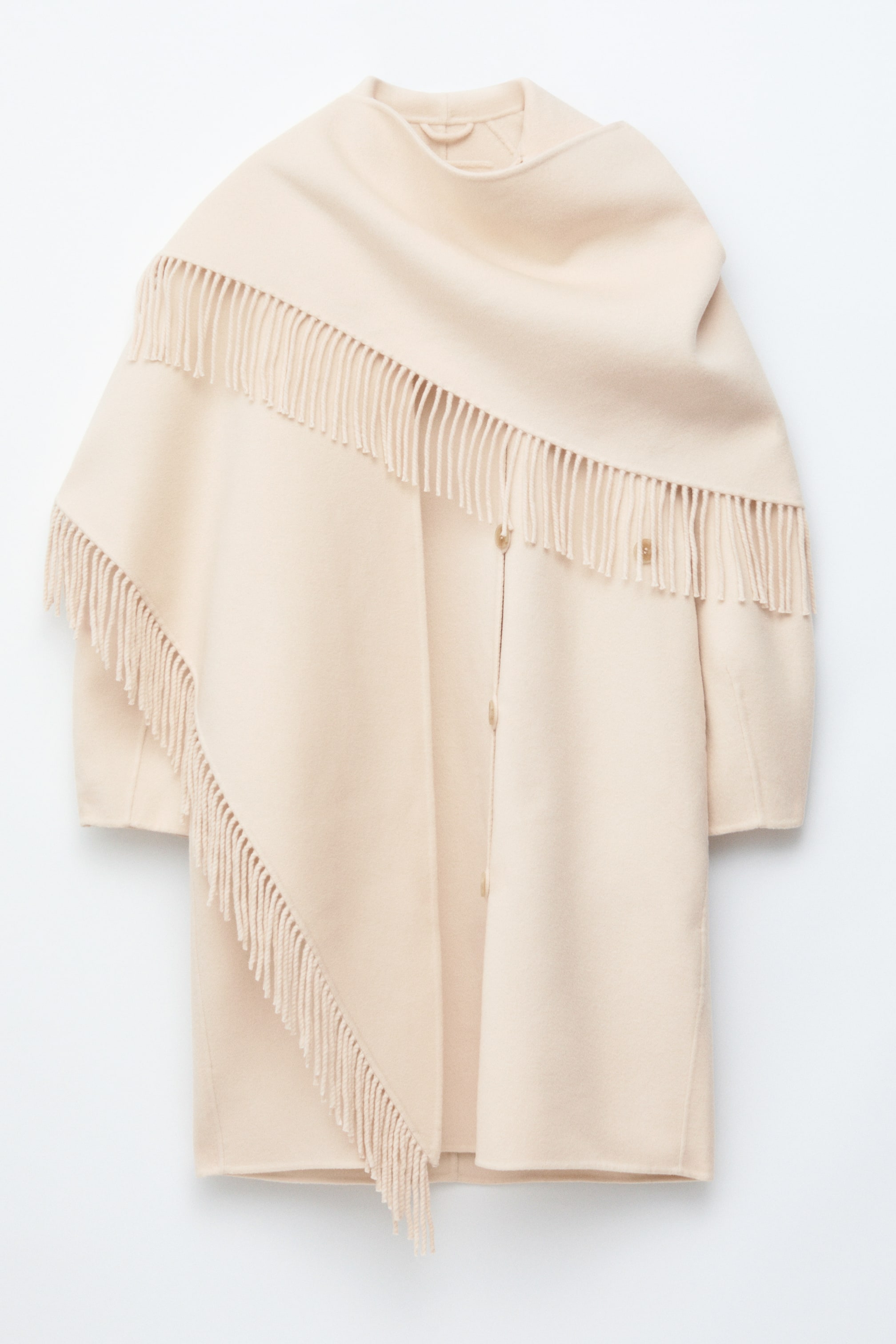 Grotere afbeelding bekijken: De crèmekleurige Fringed Shawl Wool Coat wordt frontaal gepresenteerd, met een grote sjaal die over de schouders en langs de voorkant hangt. Deze sjaal heeft lange franjes langs de diagonale randen, en het hoofddeel van de jas toont een rij lichtbruine knopen in het midden en lange mouwen.