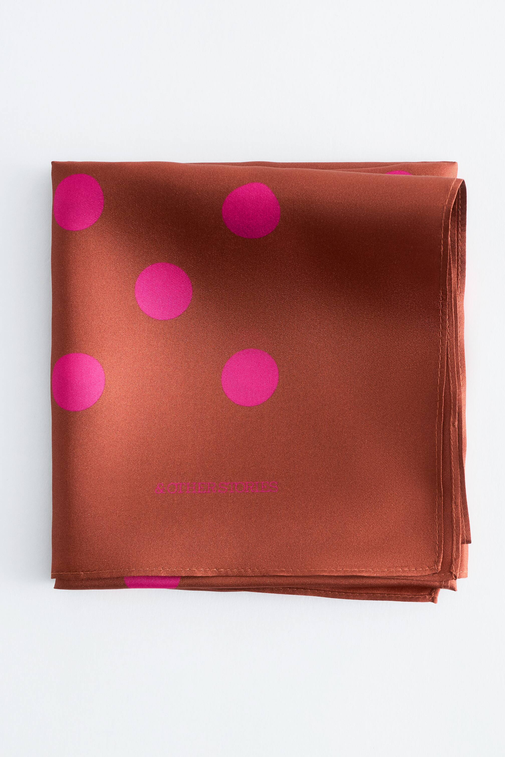 Grotere afbeelding bekijken: Een roestkleurige sjaal ligt plat, gevouwen in een vierkant, met grote fuchsia stippen en het "& OTHER STORIES" logo zichtbaar in fuchsia tekst.