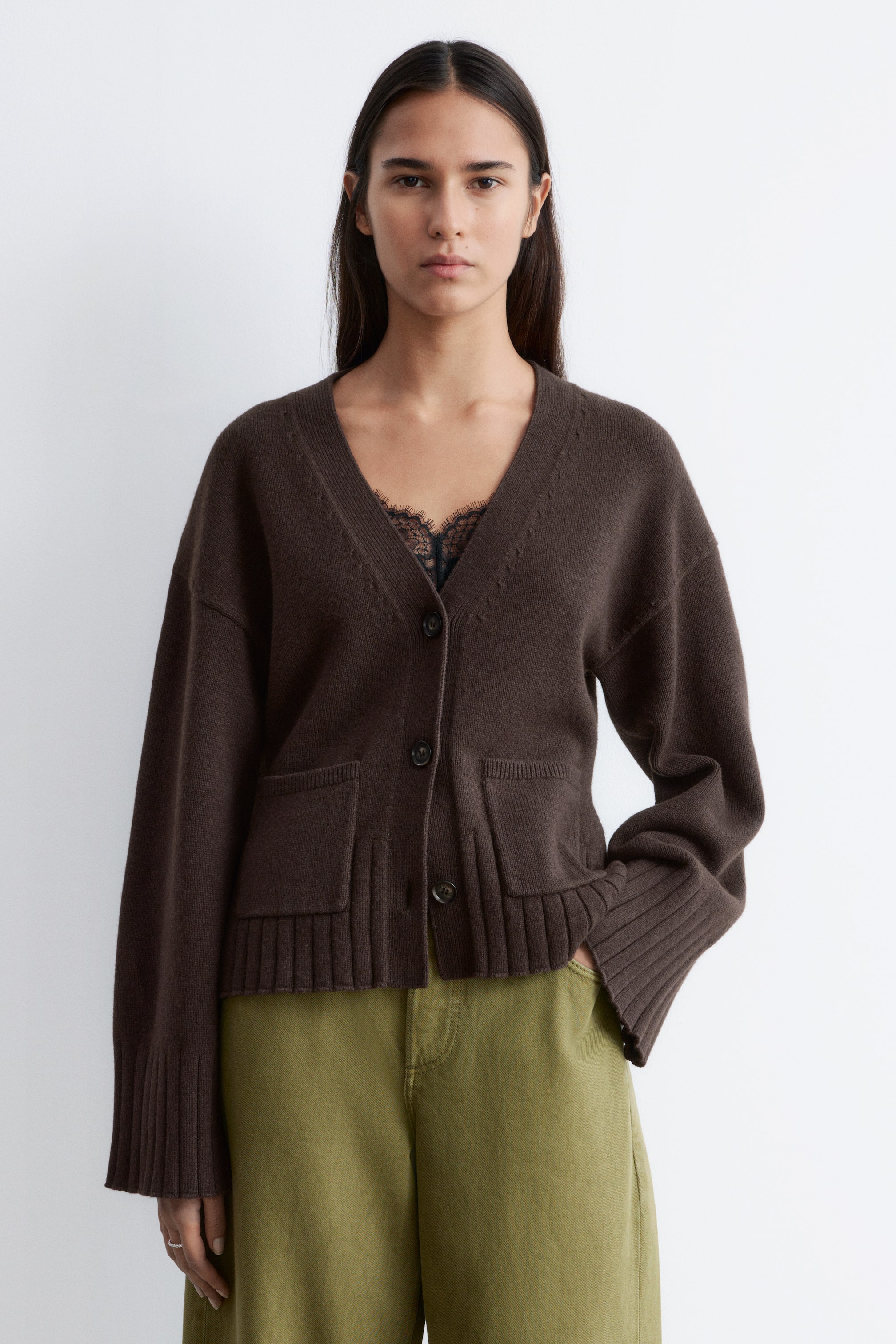 Ver imagem maior: Um cardigan castanho apresenta um decote em V e fecho frontal com botões. Apresenta dois bolsos aplicados à frente, punhos canelados e bainha canelada. O cardigan está sobreposto a um top preto com acabamento em renda.