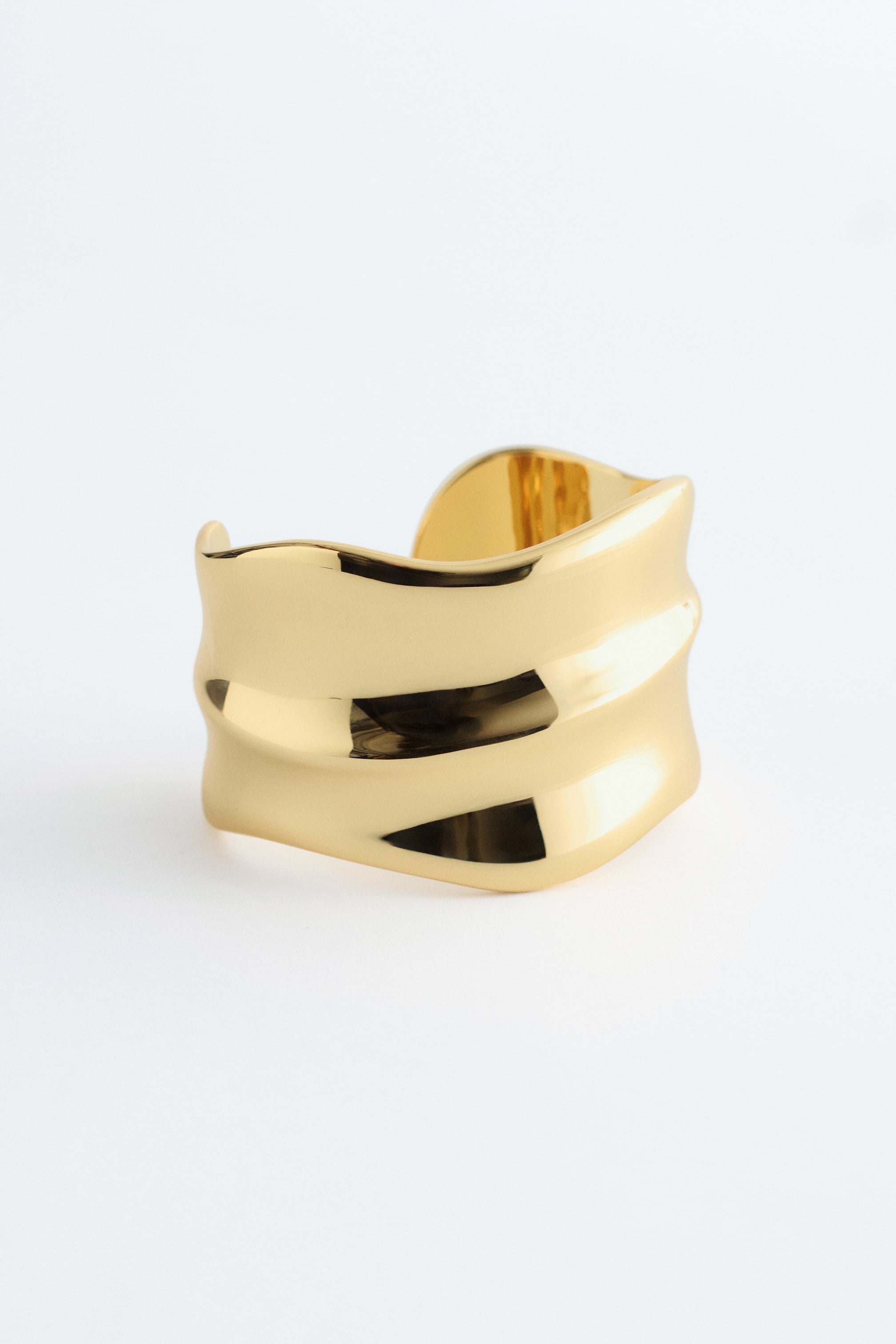 Grotere afbeelding bekijken: De Wavy Cuff Armband is frontaal gericht en heeft een brede, glanzende goudkleurige band die doorlopende, golvende bewegingen langs het oppervlak vormt.