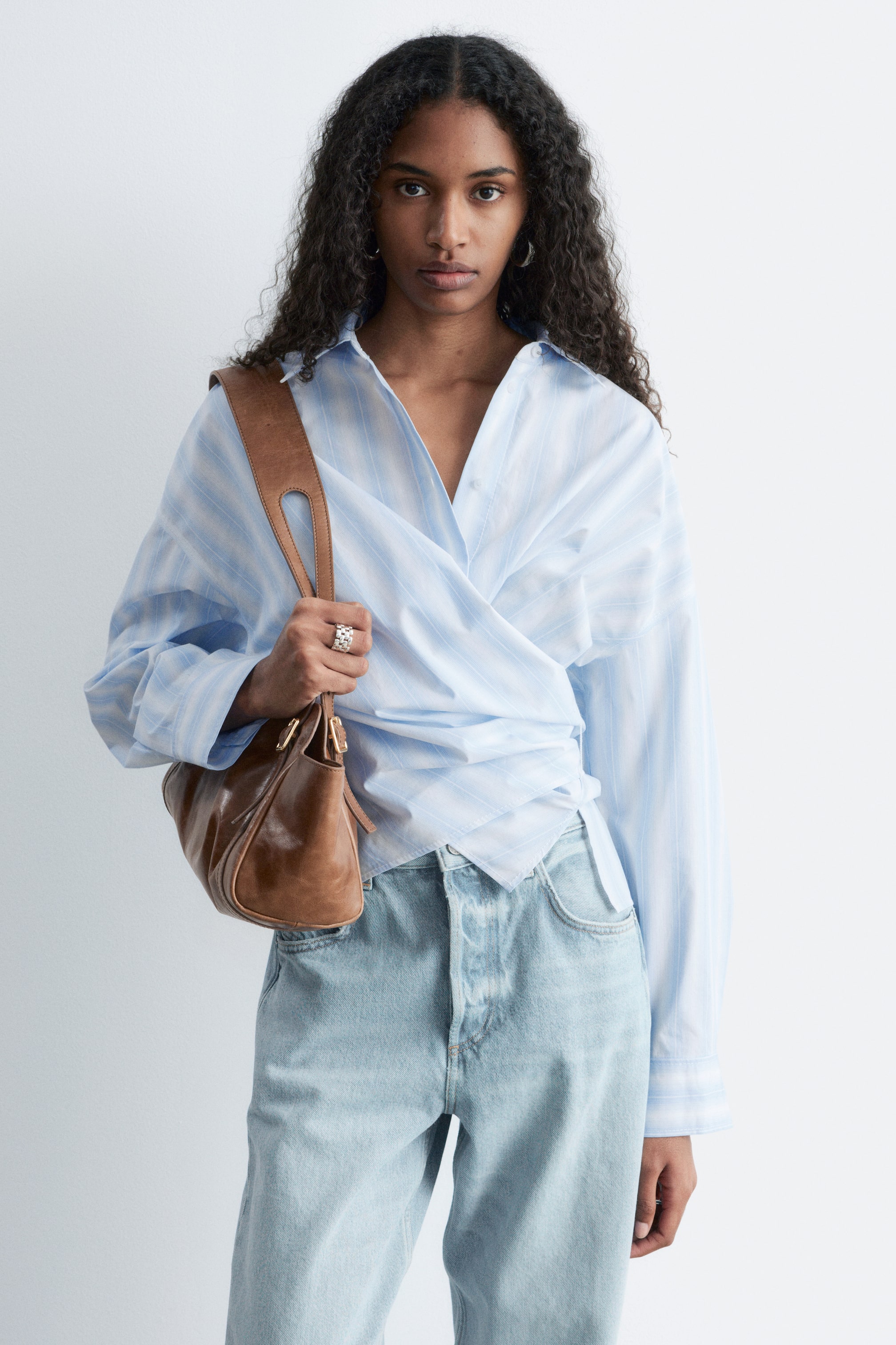 Zobraziť väčší obrázok: Modelka má na sebe svetlomodrú pruhovanú zavinovaciu košeľu s dlhými rukávmi a špicatým golierom. Predná časť košele je zavinovacia a viaže sa v páse, čím vytvára skrátenú siluetu. Modelka má tiež oblečené svetlé džínsy a nesie hnedú kabelku na rameno.