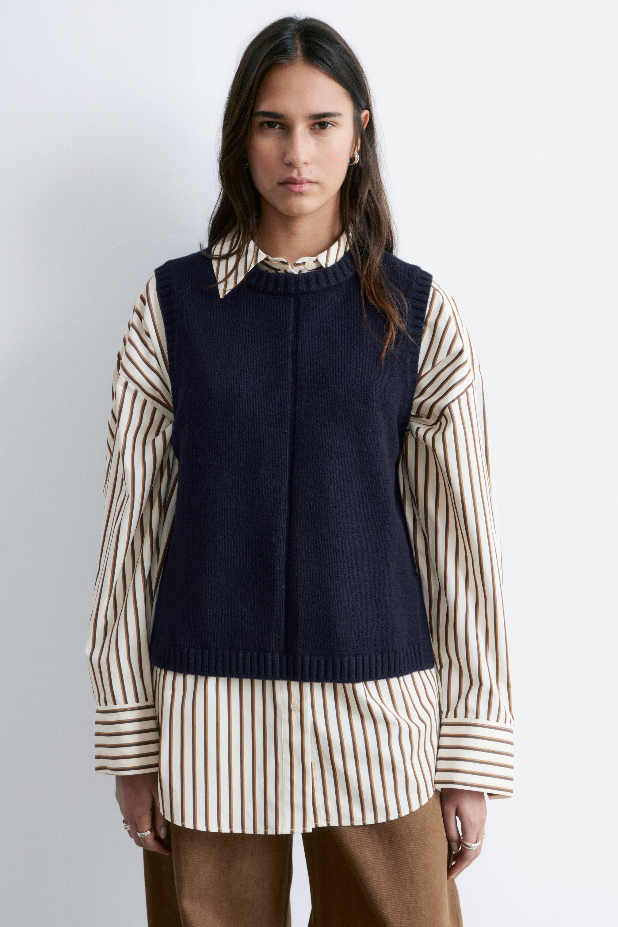 Ver imagen más grande: Un chaleco de punto azul marino se presenta desde el frente, superpuesto sobre una camisa a rayas. Presenta un cuello redondo acanalado, una costura vertical central que recorre la parte delantera y un dobladillo acanalado.